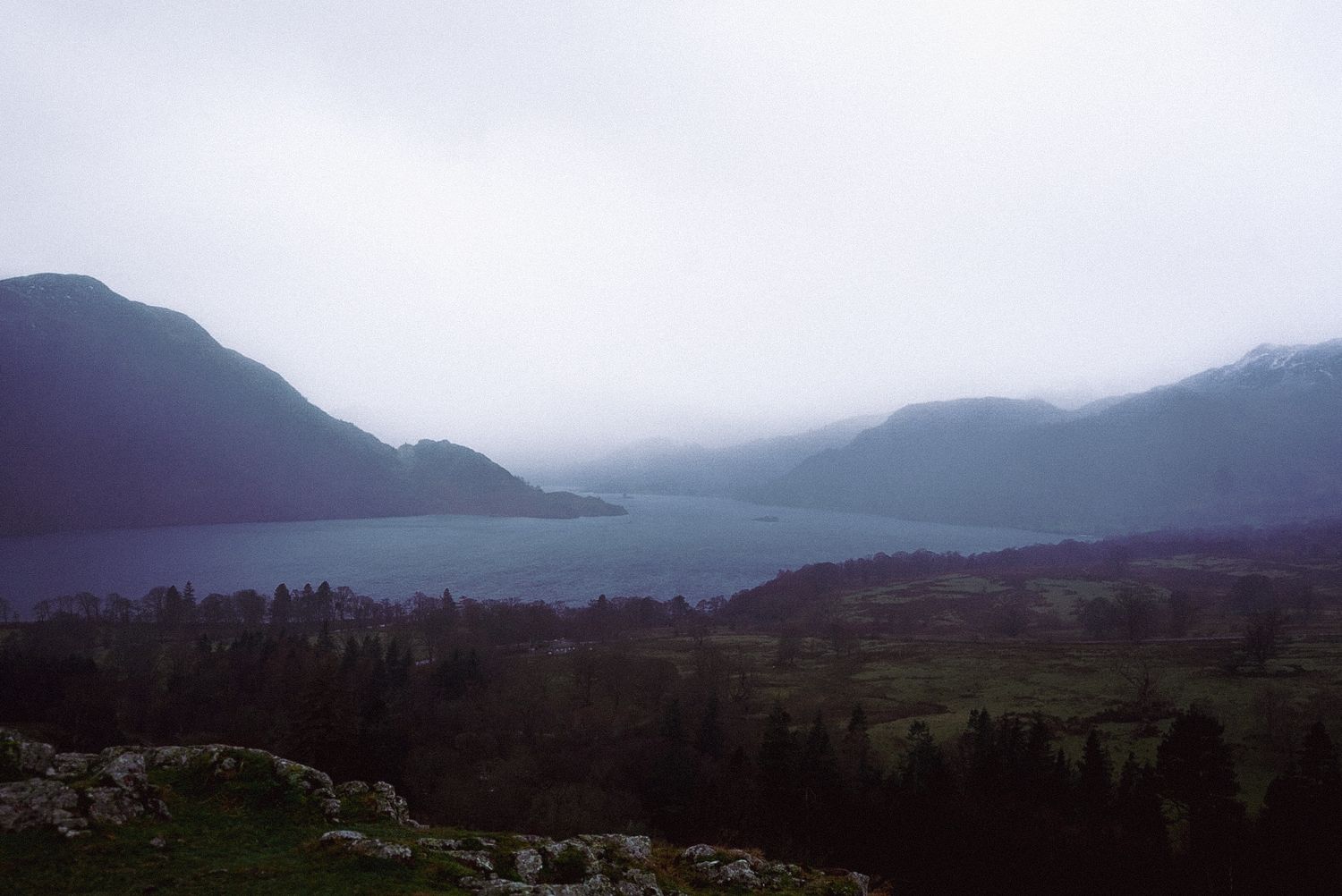 Dramatic misty lake valley surrounded by dark mountains and evergreen forest.