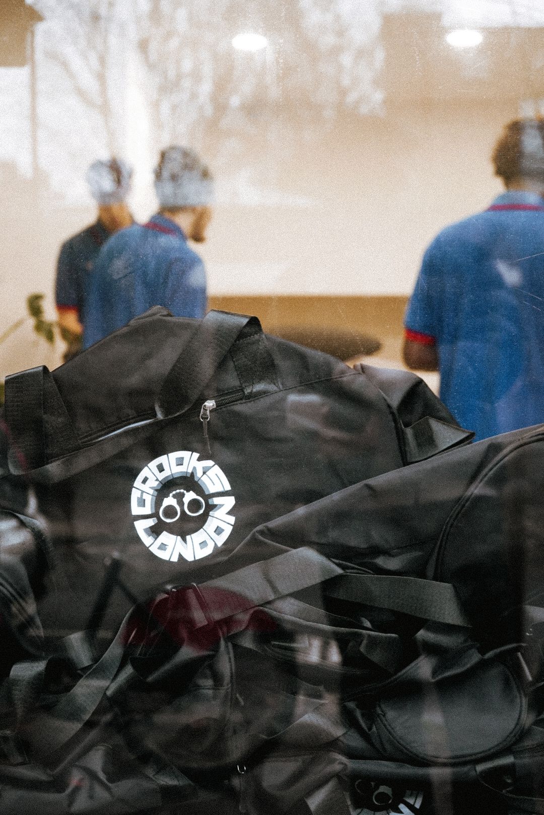 A black Crooks London duffel bag sits in the foreground with people reflected in a window behind.