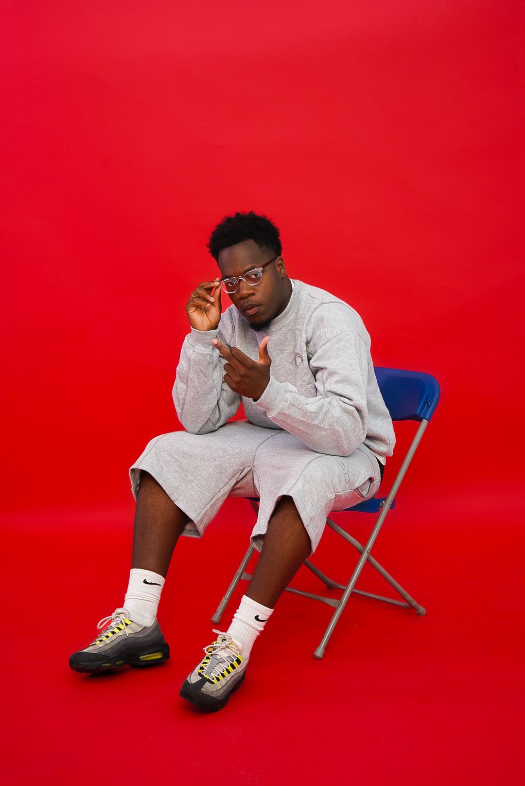 Young man in grey sweatsuit adjusts glasses while sitting on a blue folding chair against red background.