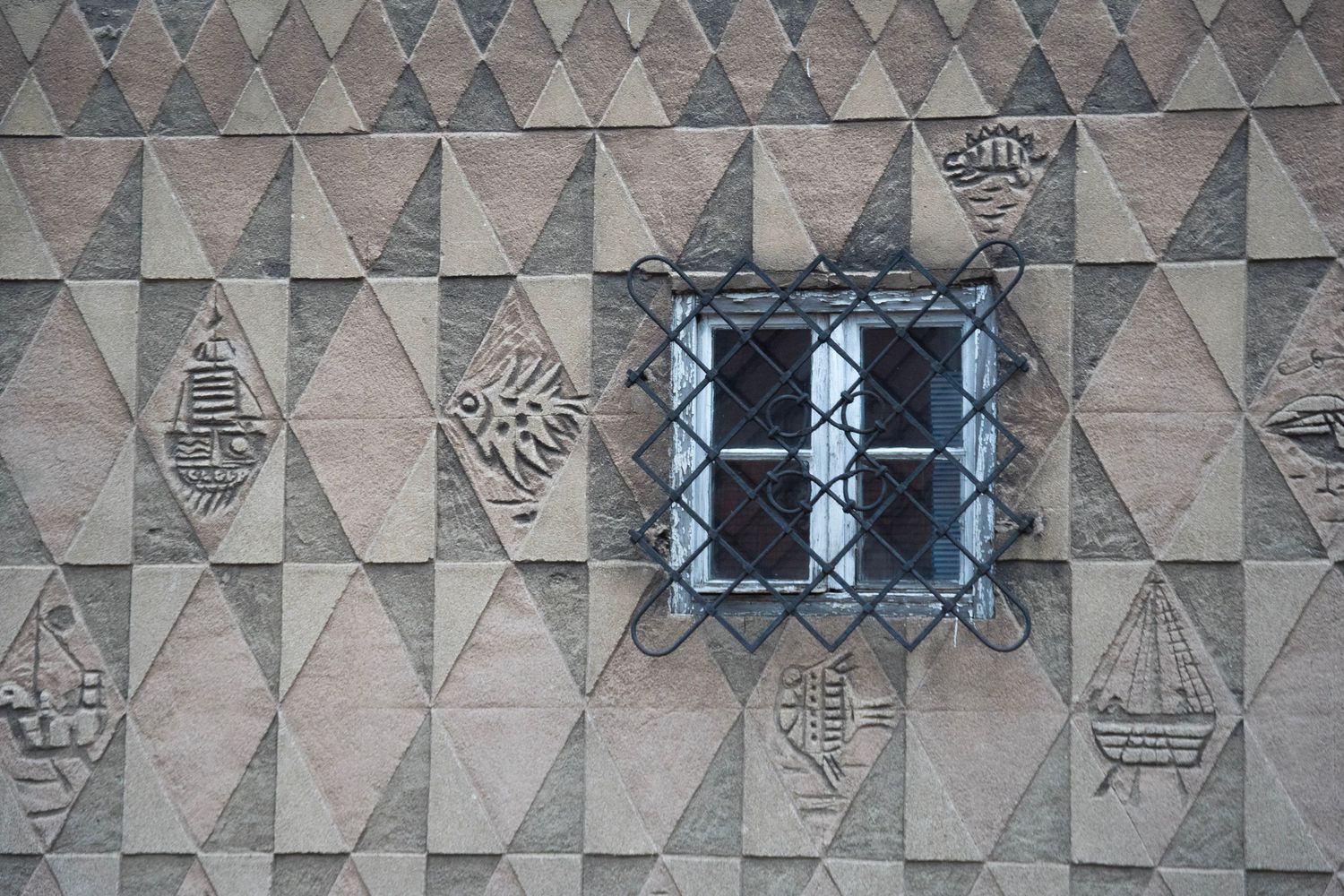 Stone wall with diamond-pattern reliefs featuring fish and ship carvings, centered around a barred window.