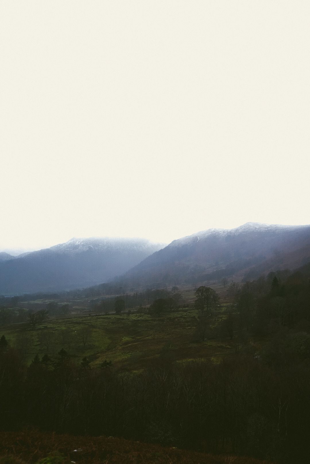 Overcast mountain valley landscape with layered hills and sparse trees in mist.