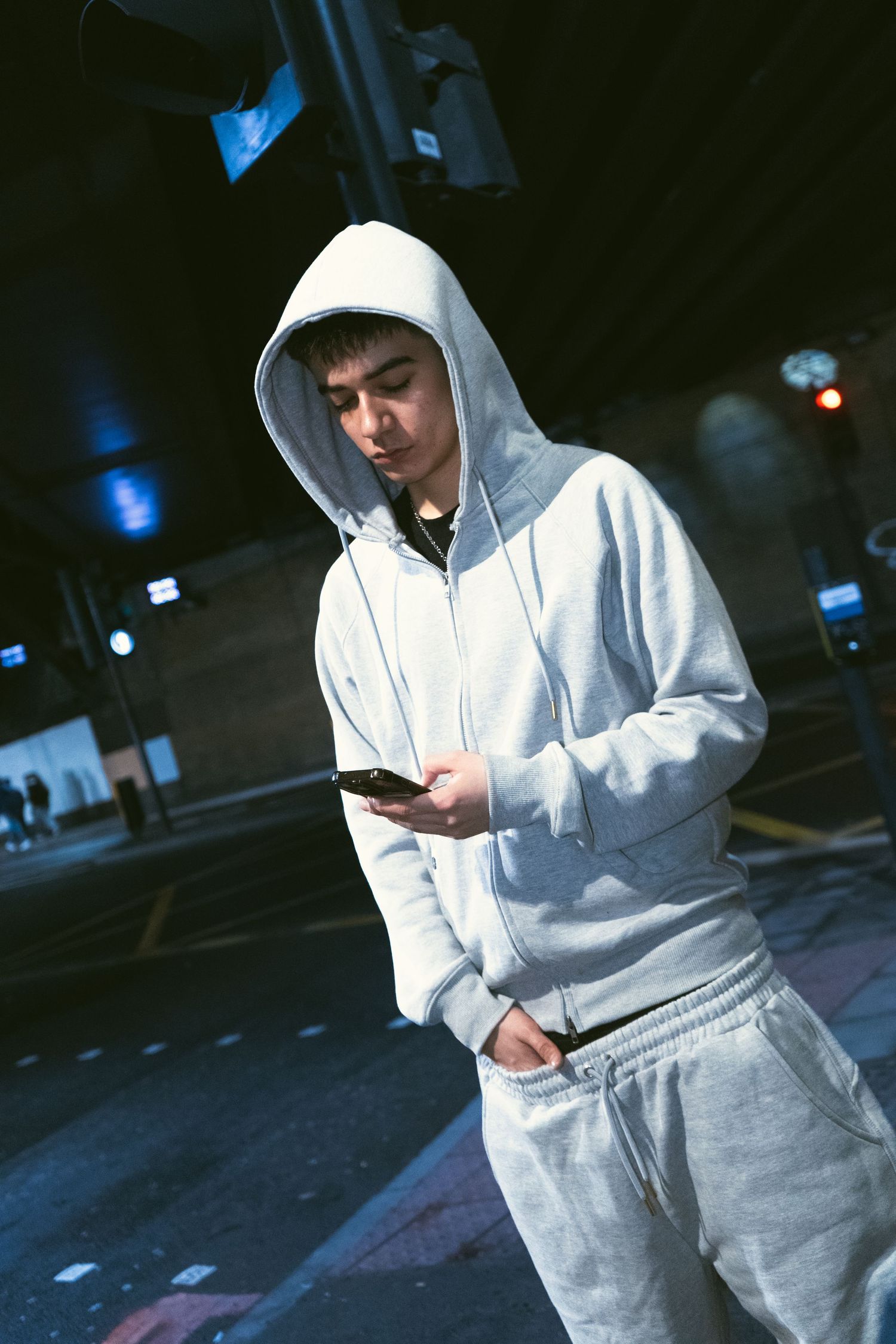 Young man in grey hoodie and sweatpants checking phone on urban street at night.