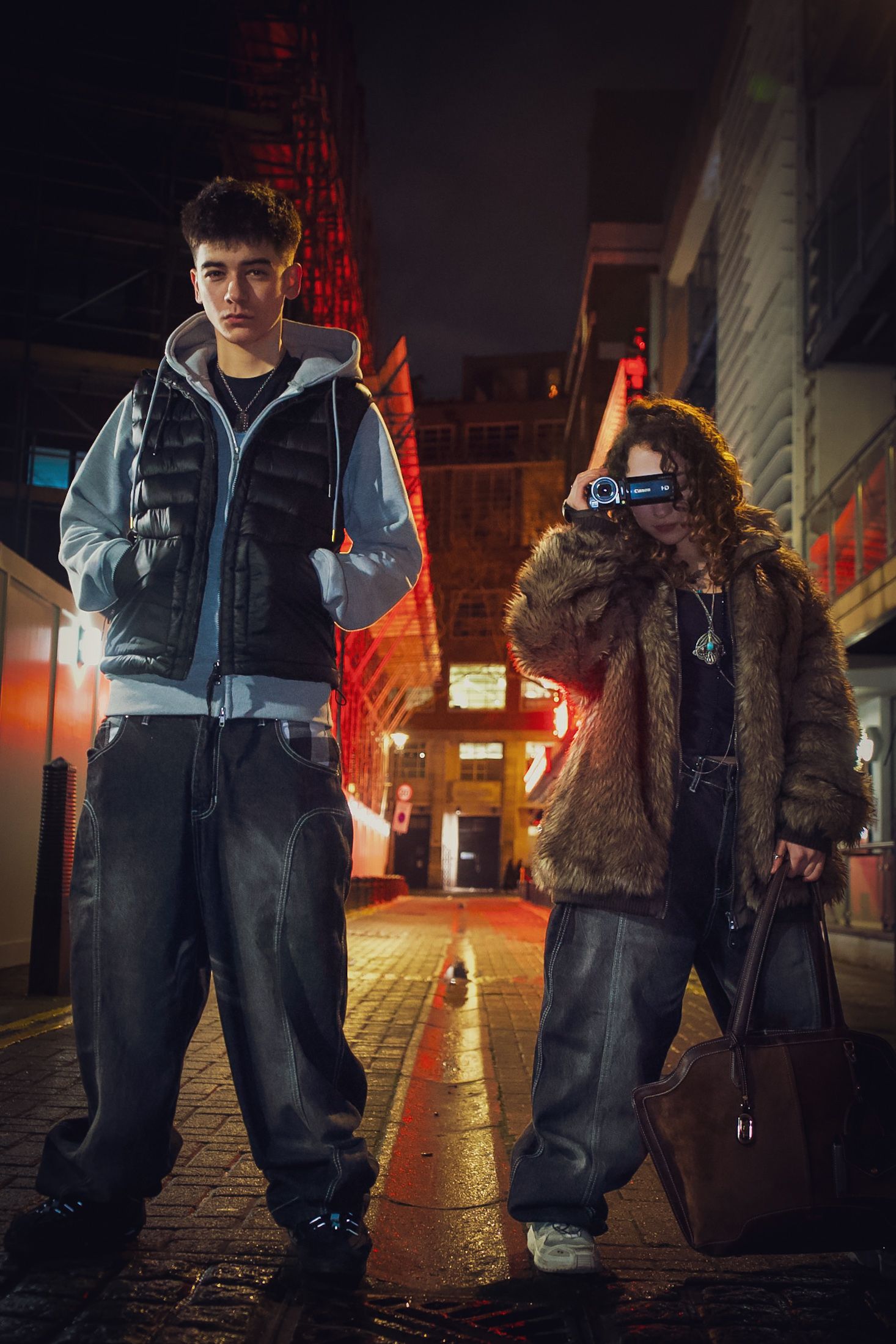 Two young people in streetwear posing on a wet cobblestone street at night with red neon lights.