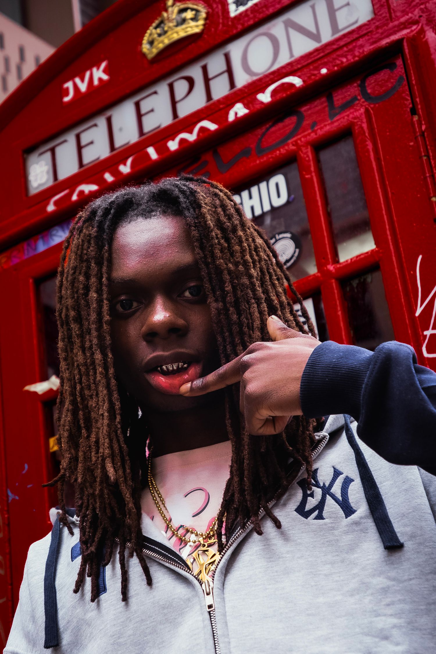 Young man with dreadlocks and gold grills poses in front of a red London telephone booth.