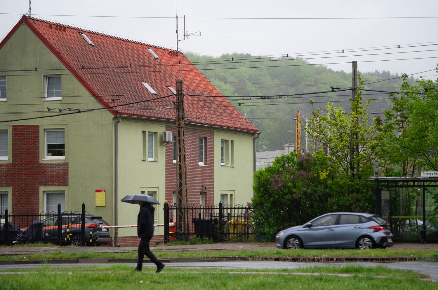 Person walking with umbrella on rainy day near residential buildings with red roofs and parked silver car.