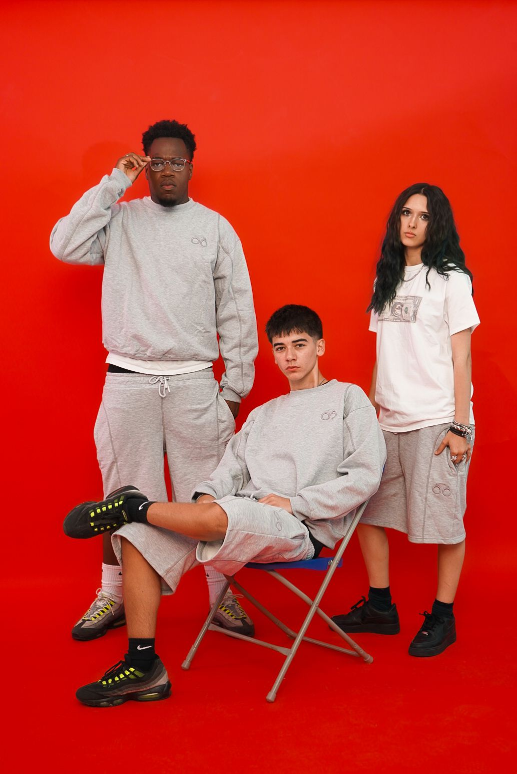 Three diverse young people in matching grey streetwear sets pose together against a vibrant red background.