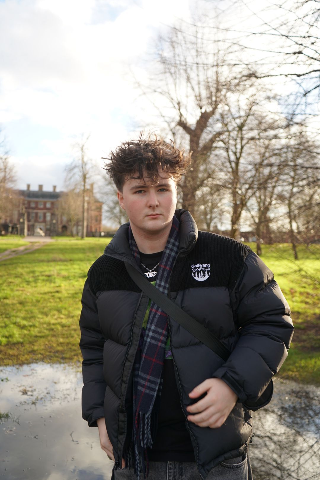 Young man in black puffer jacket and plaid scarf stands in a sunny green park setting.