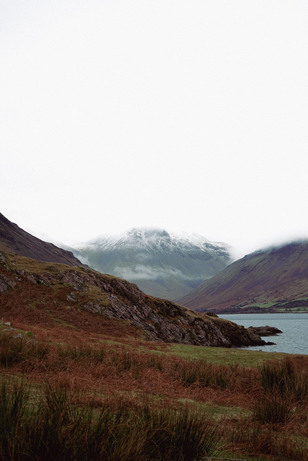 A snow-capped mountain shrouded in mist rises above a calm lake surrounded by rugged terrain.