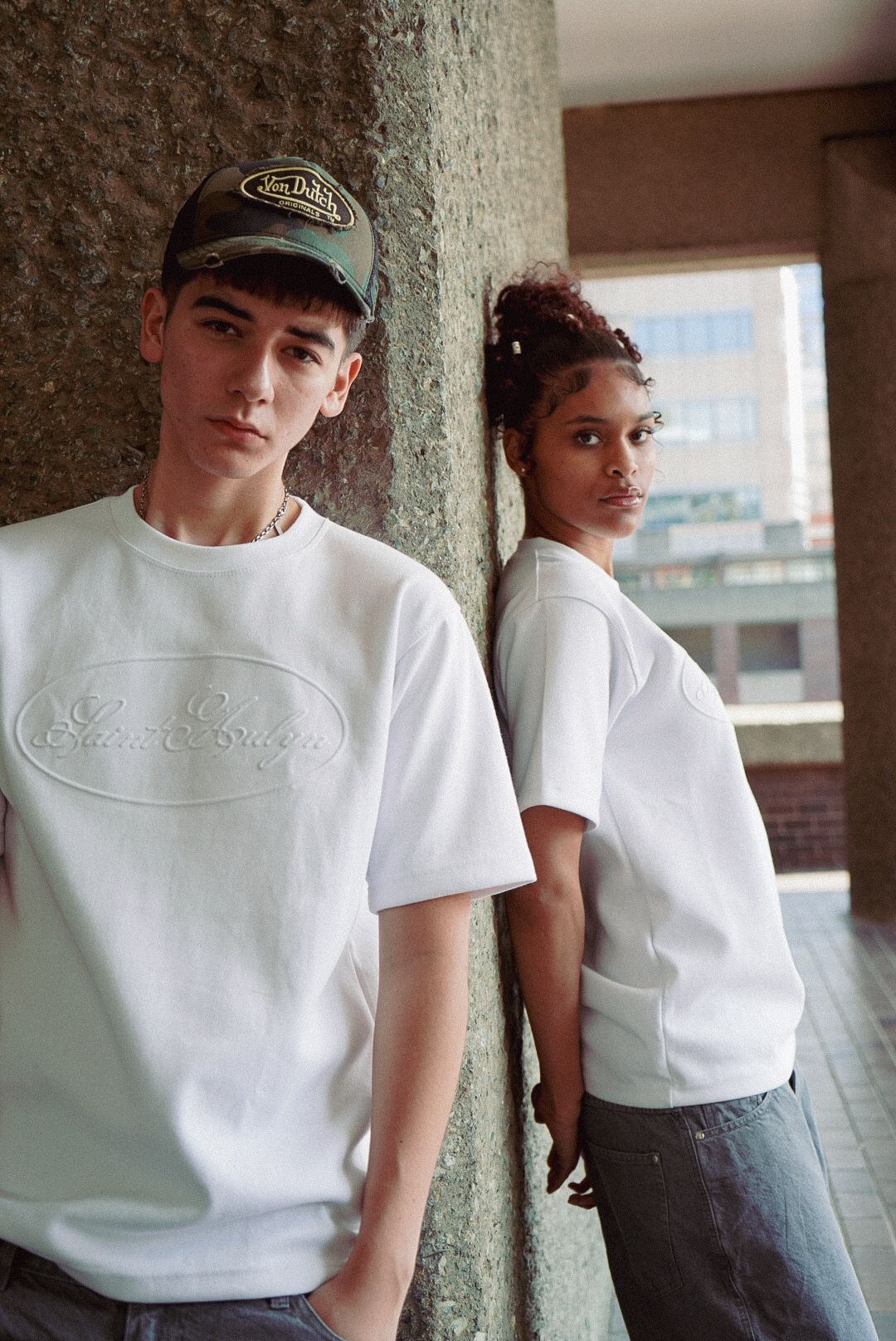 Two young models wearing matching white oversized logo t-shirts leaning against concrete pillar.