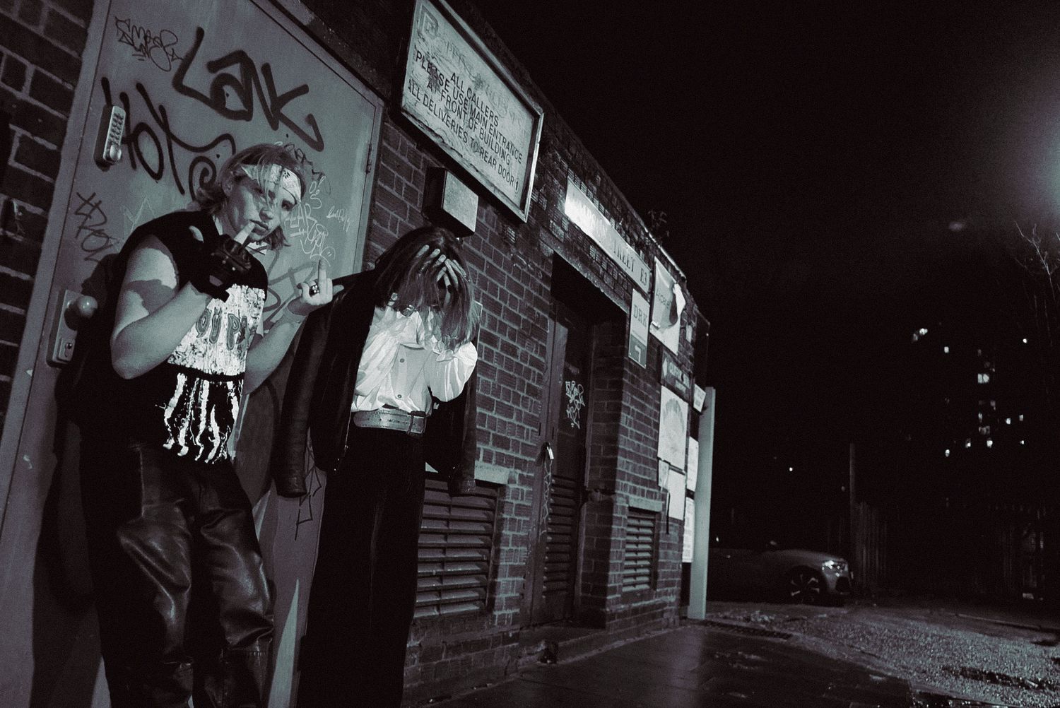 Black and white night photo of people standing outside a graffiti-covered brick building.