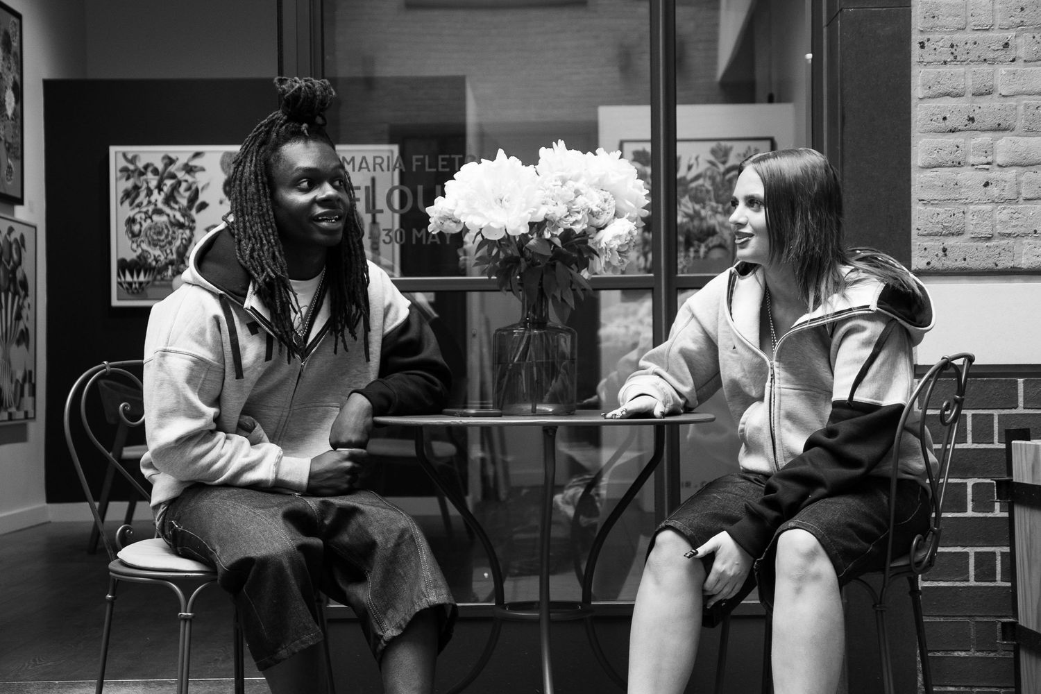 Two people sitting and conversing at a glass table in a stylish cafe, black and white photo with flowers in background.