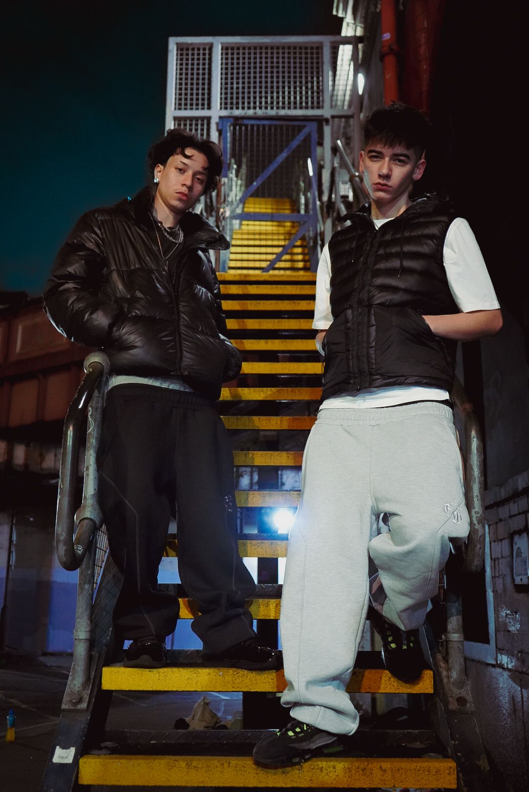 Two young men in black puffer vests stand on yellow industrial stairs in an urban night setting.