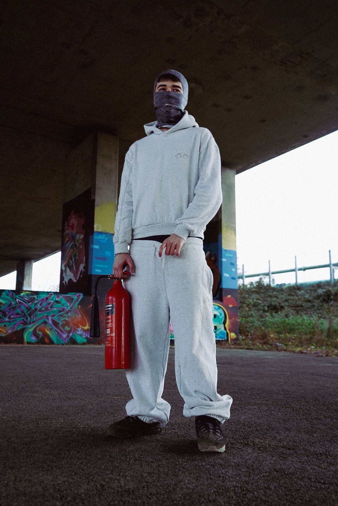 Masked figure in grey hoodie and sweatpants holds red fire extinguisher under bridge with graffiti.
