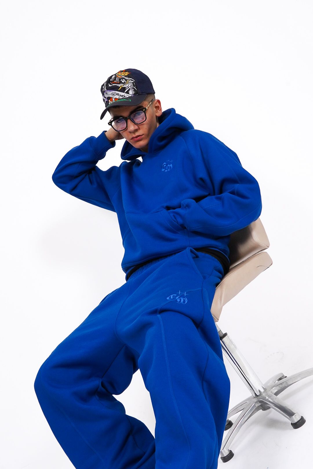 Young man in bright blue oversized hoodie and matching joggers sitting on chair in white studio.