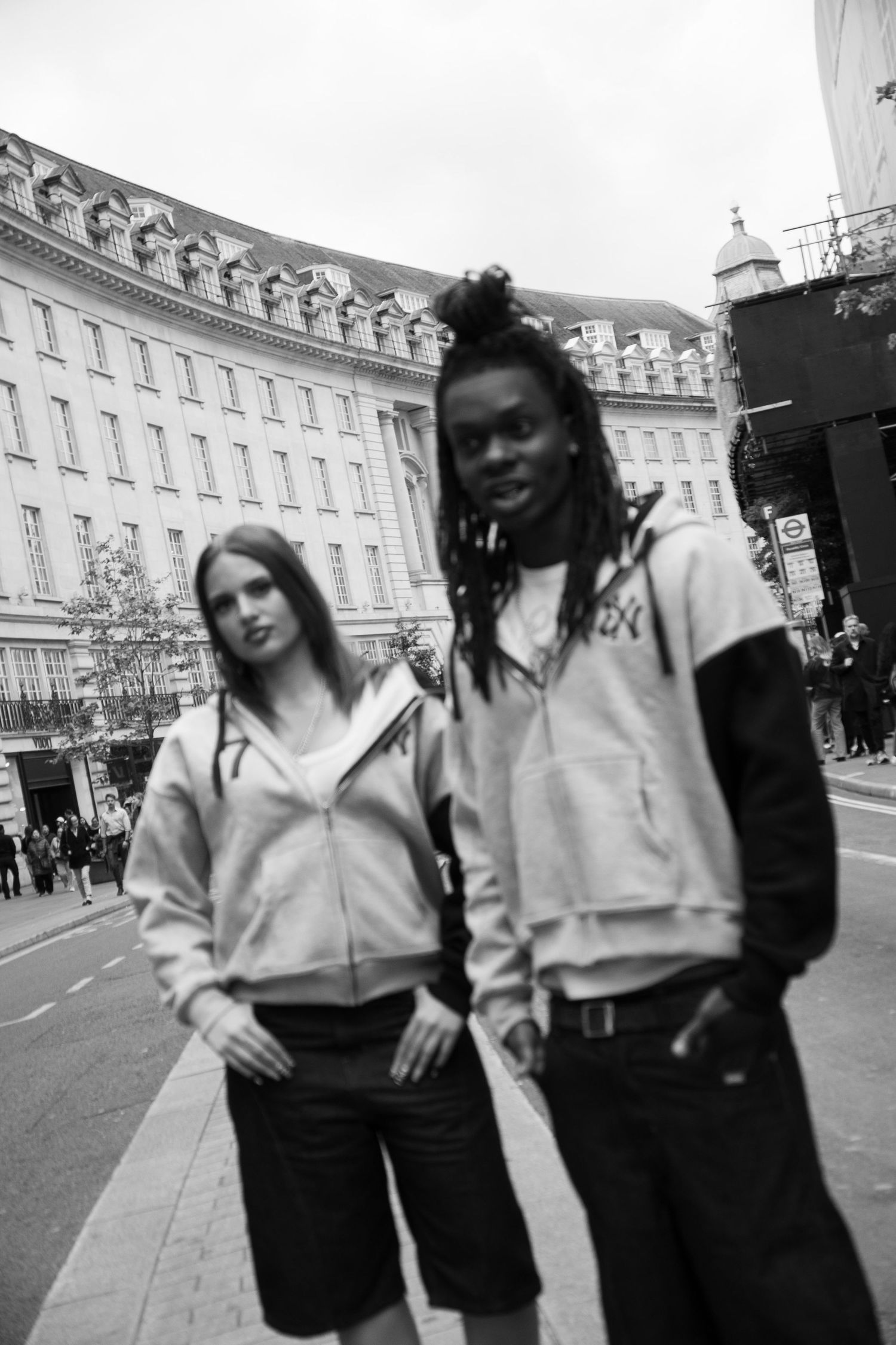 Black and white photo of two young people in hoodies on a London street with a curved building in the background.