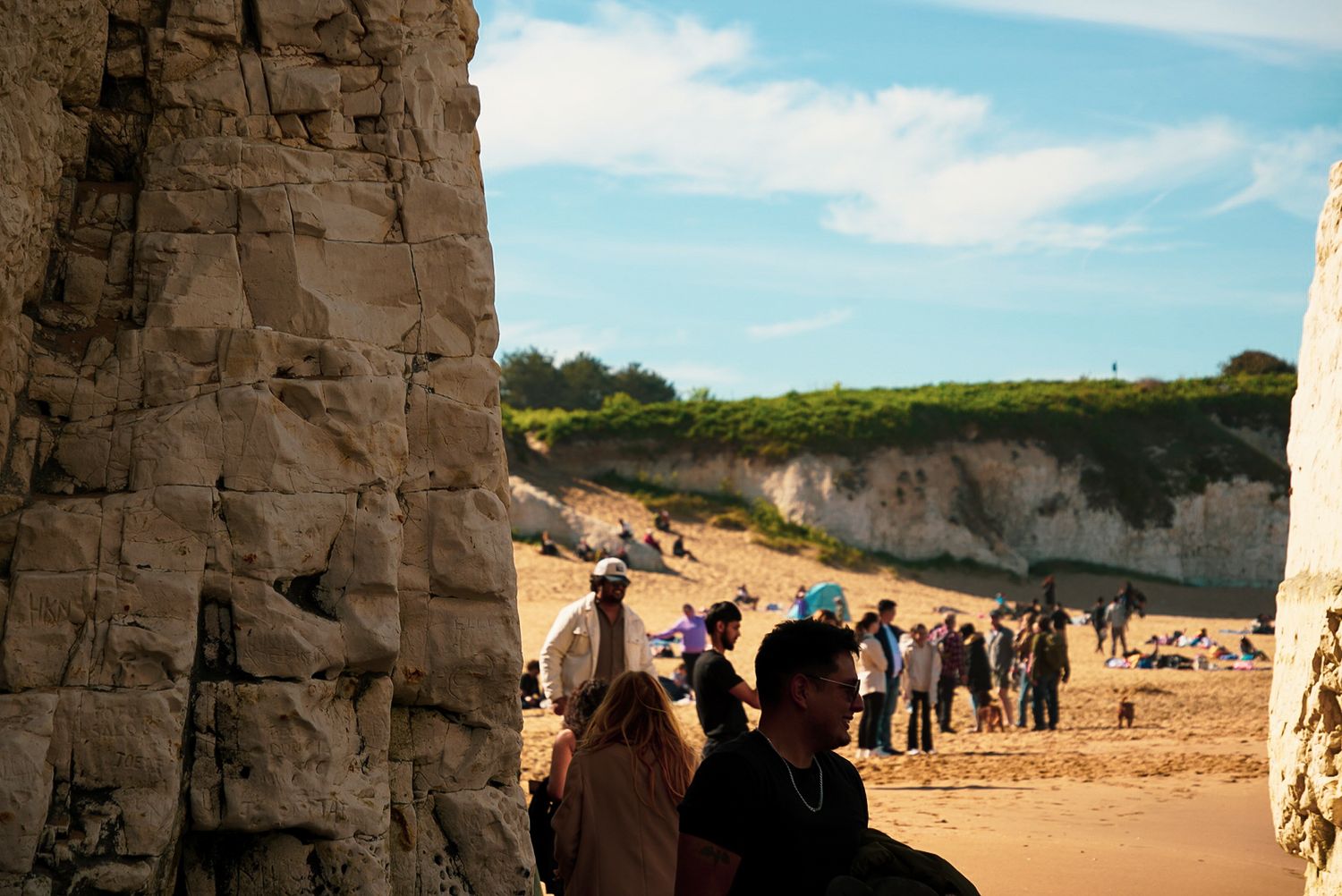 Tourists gather on a sandy beach near white chalk cliffs under a blue sky on a sunny day.