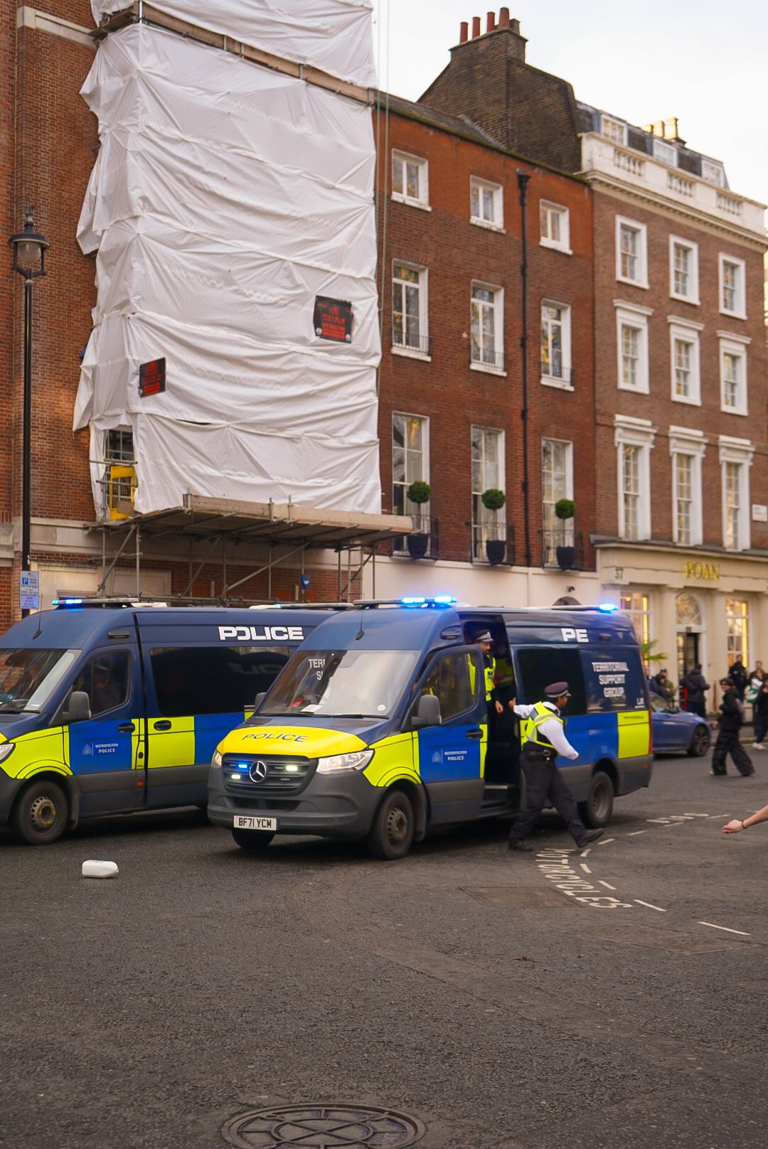 Blue Metropolitan Police vans with yellow markings parked on a London street beside a scaffolded brick building.