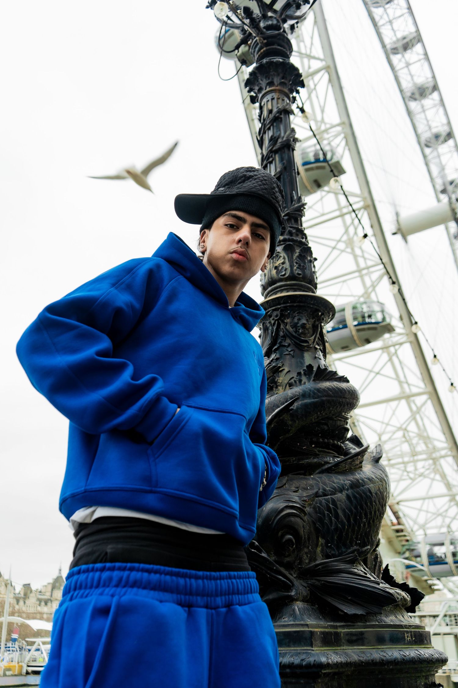 Young person in blue tracksuit leaning on ornate lamppost with London Eye in background.
