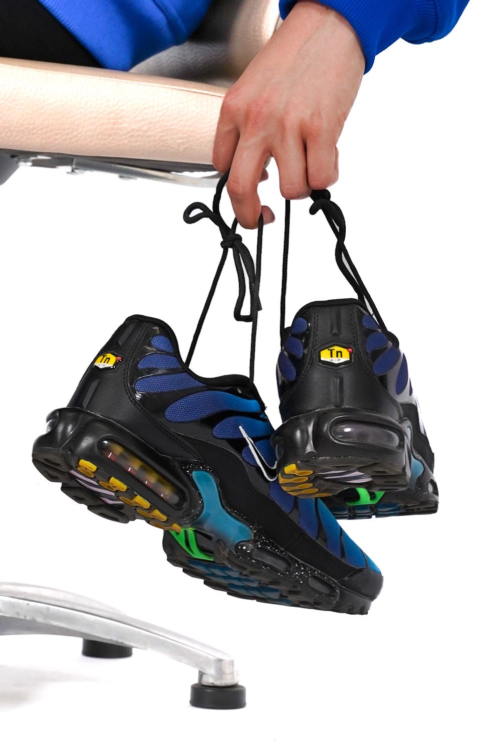 Close-up of hands holding Nike Air Max Plus sneakers by laces over chair in blue sleeve.