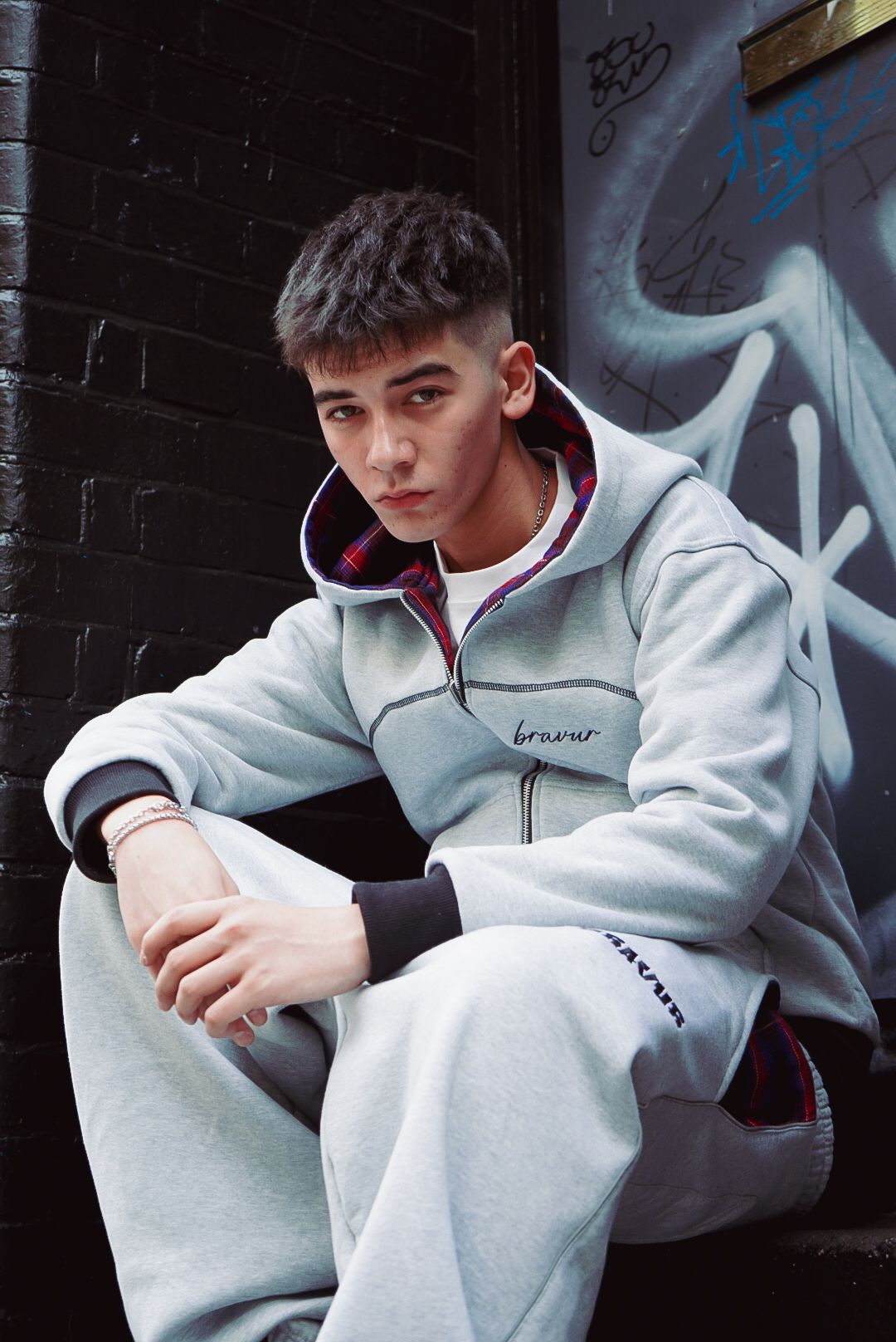 Young man in grey Bravur zip-up hoodie and sweatpants sits relaxed against a graffiti brick wall.