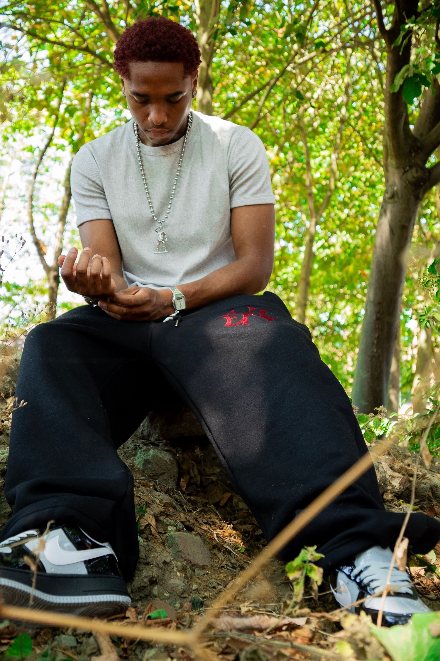 Young man in white tee and black sweats sits outdoors among trees, looking down at his hands.