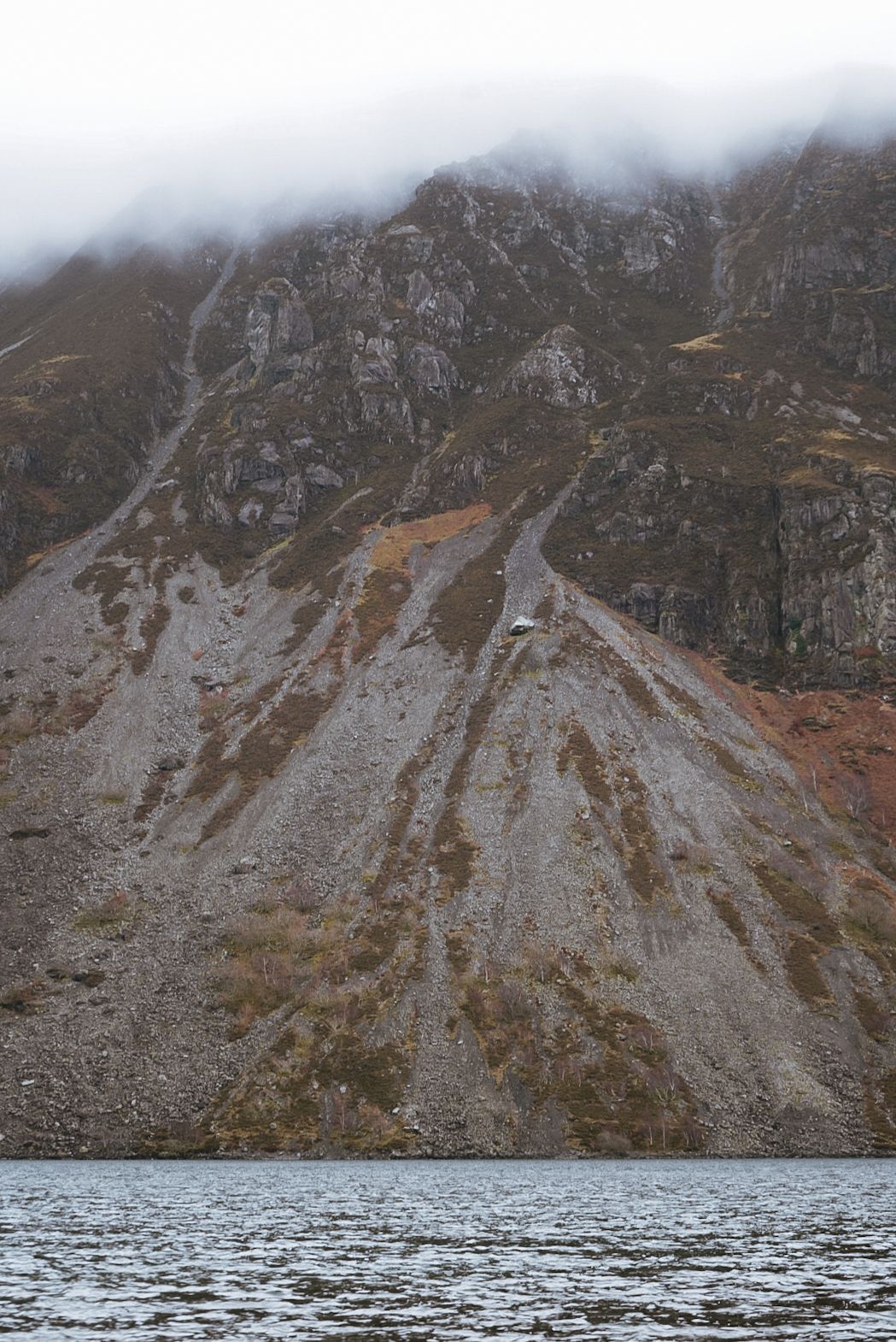 A steep rocky mountain scree slope descends to a dark lake under a cloudy misty sky.