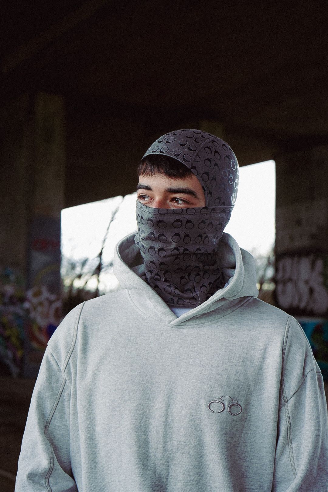 Young man wearing grey balaclava and light grey hoodie with small logo standing in urban setting.