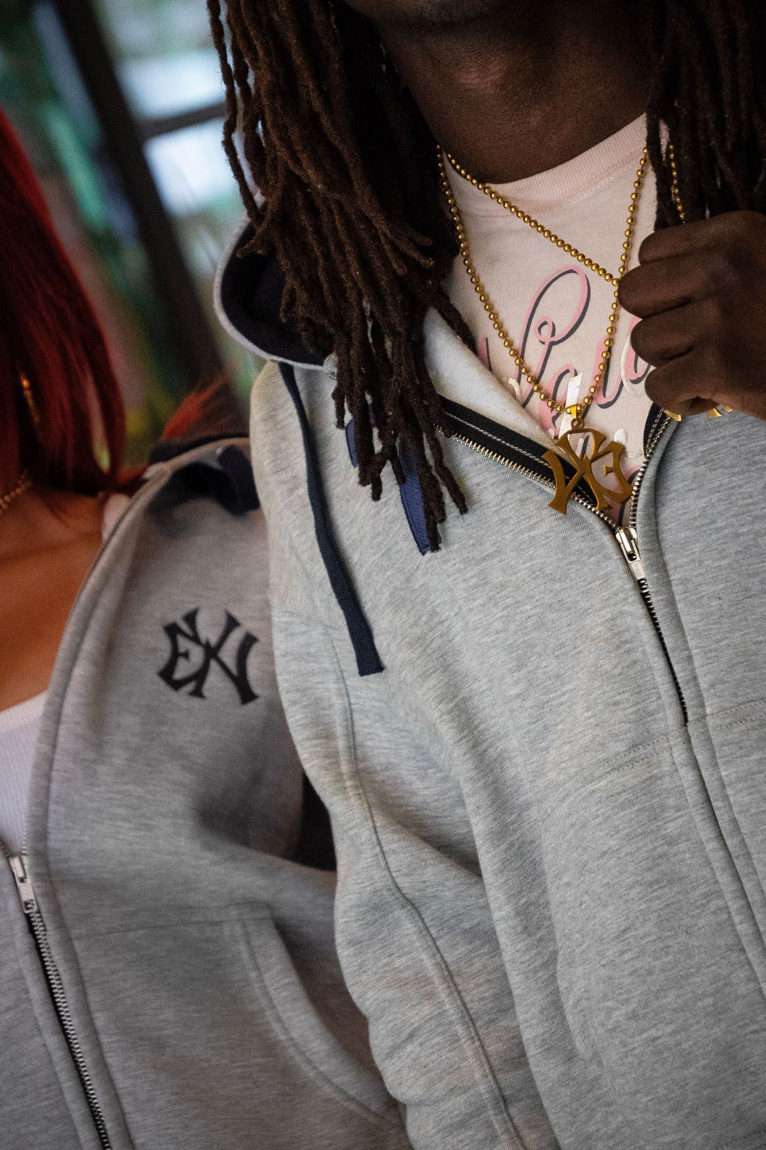 Man with dreadlocks wearing a gray NY Yankees hoodie and gold chain necklaces stands next to a woman with red hair.