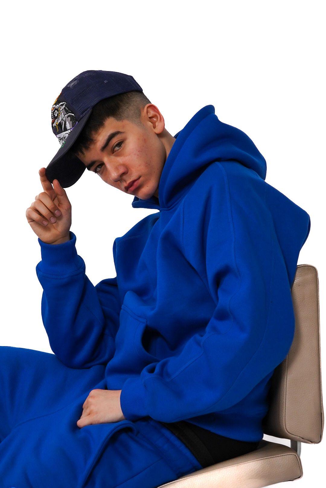 Young man in oversized royal blue hoodie sitting and adjusting navy cap on white background.