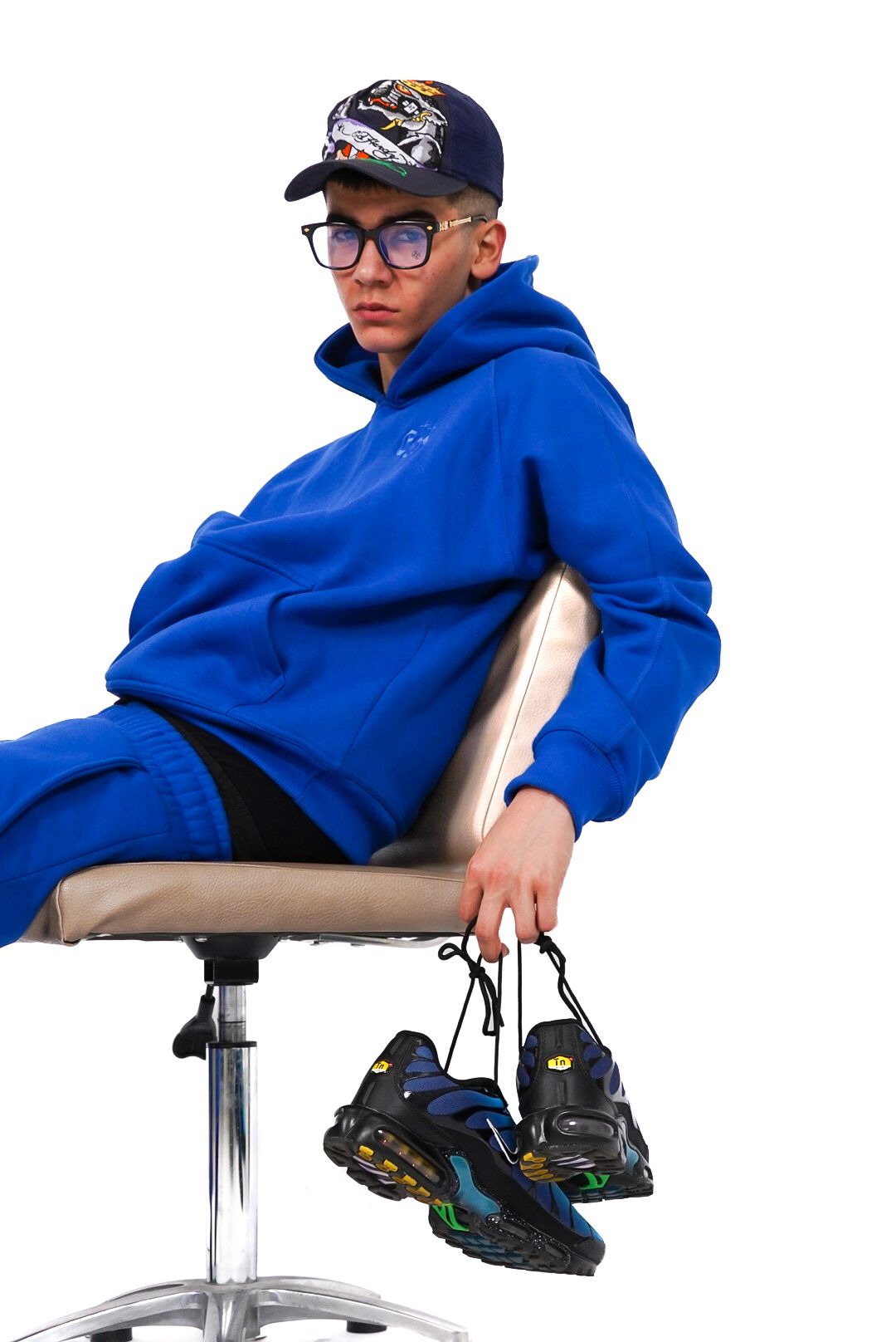 Young man in blue hoodie sitting on chair holding Nike Air Max Plus sneakers, wearing glasses and graphic cap.