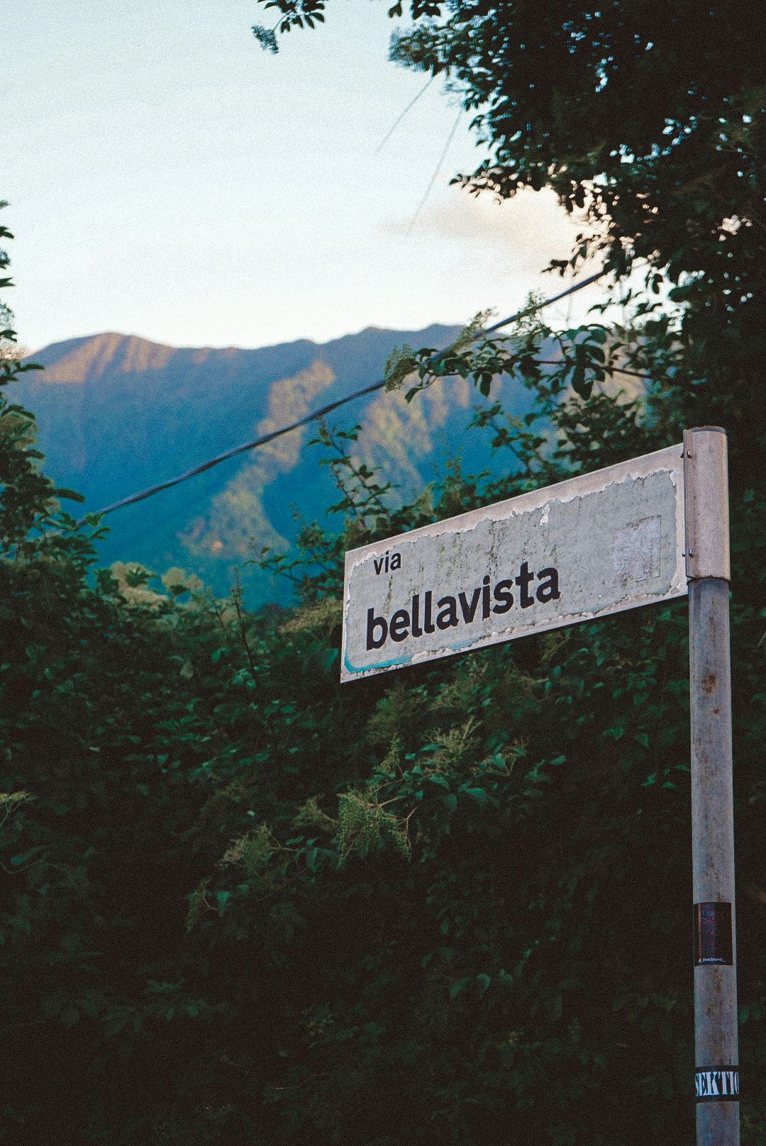 Street sign reading Via Bellavista mounted on pole surrounded by lush green trees with mountain backdrop.