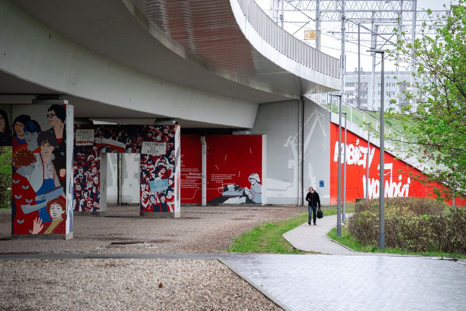 Urban underpass with colorful murals and red graffiti walls, surrounded by green trees and a pedestrian walkway.