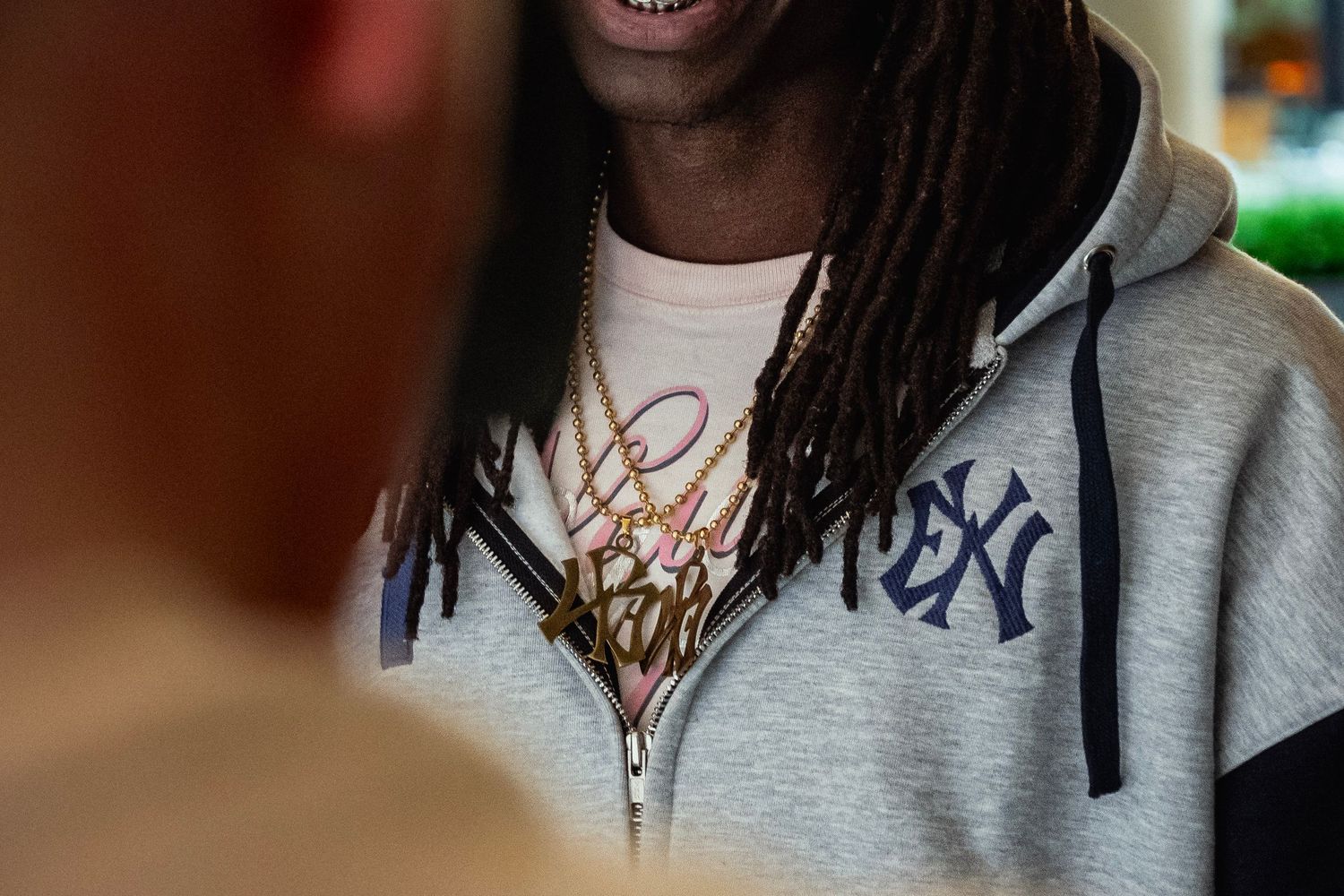 Young man with dreadlocks wearing a gray NY Yankees hoodie and gold chain necklaces.