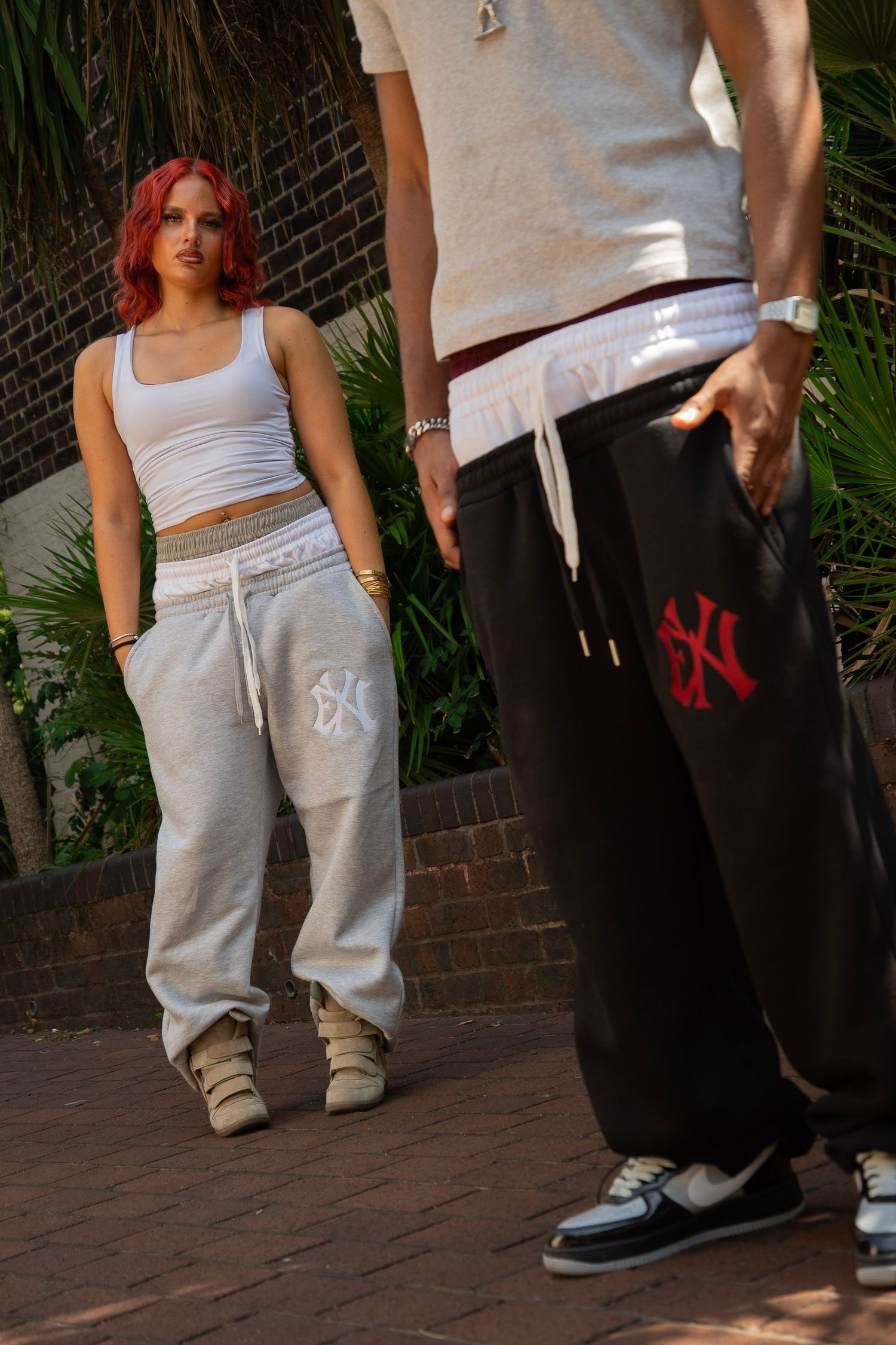 Two people wearing matching NY logo gray and black sweatpants, styled with a white crop top and beige tee outdoors.