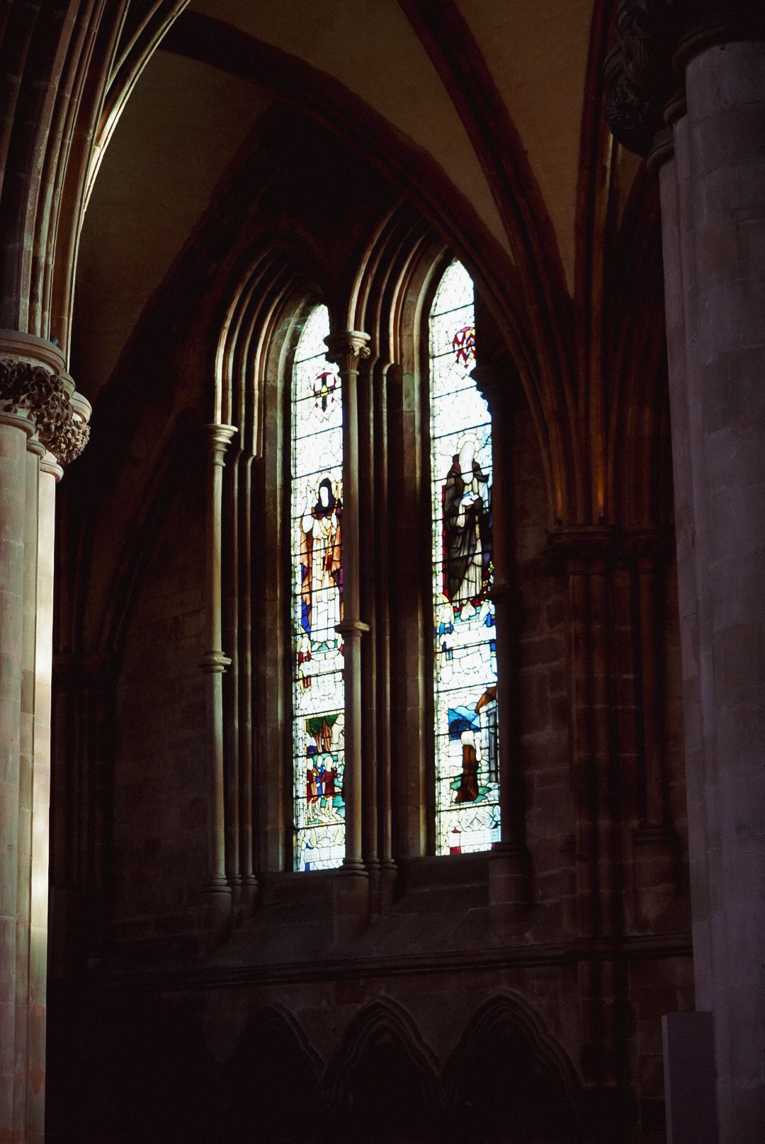 Two tall stained glass windows with colorful biblical scenes inside a dark Gothic cathedral.