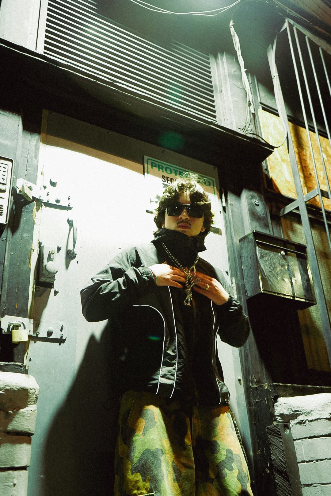Stylish person in sunglasses, black jacket, and camo pants posing in a sunlit urban alleyway.