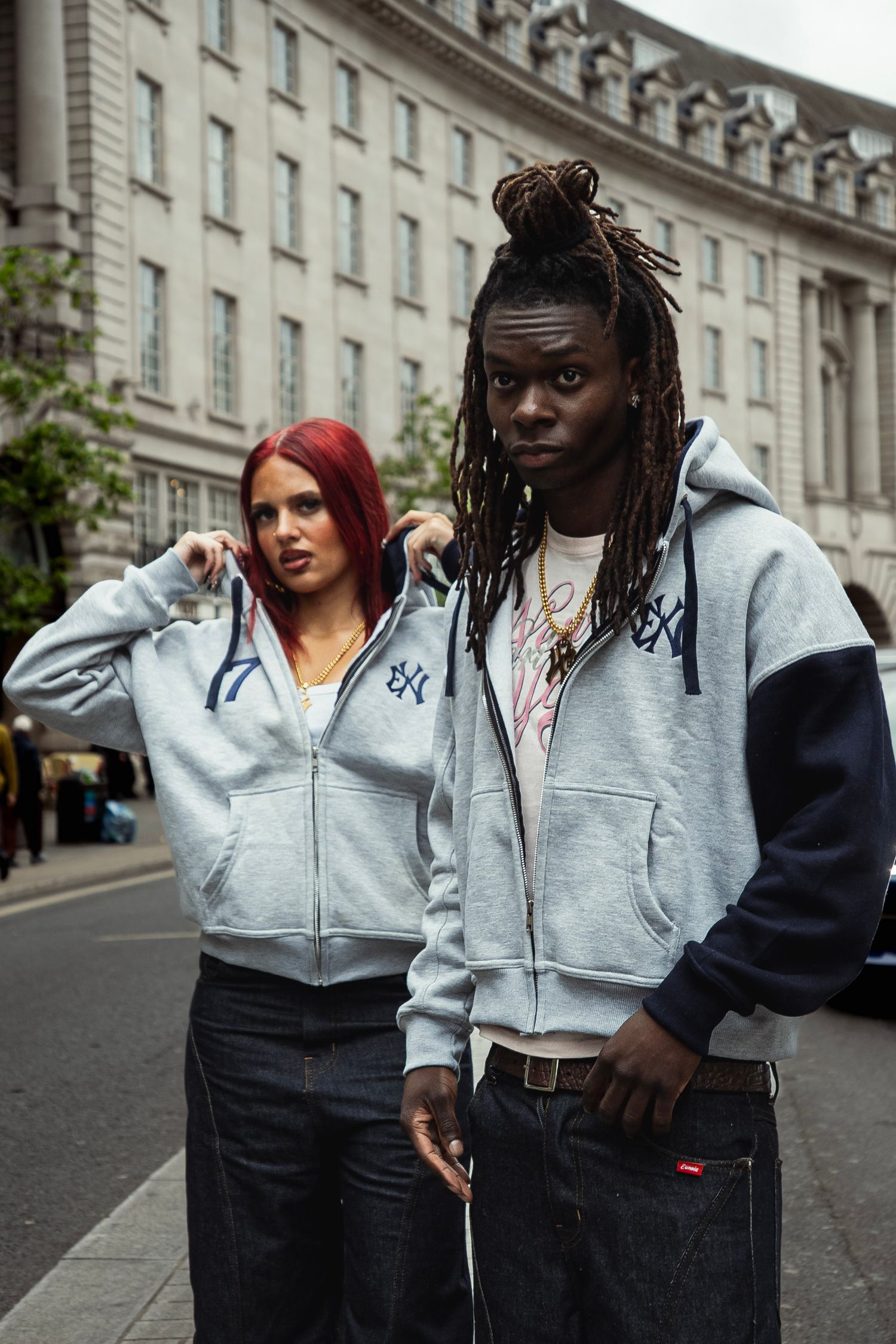 Two models in matching grey zip-up hoodies with navy accents pose on a London street.