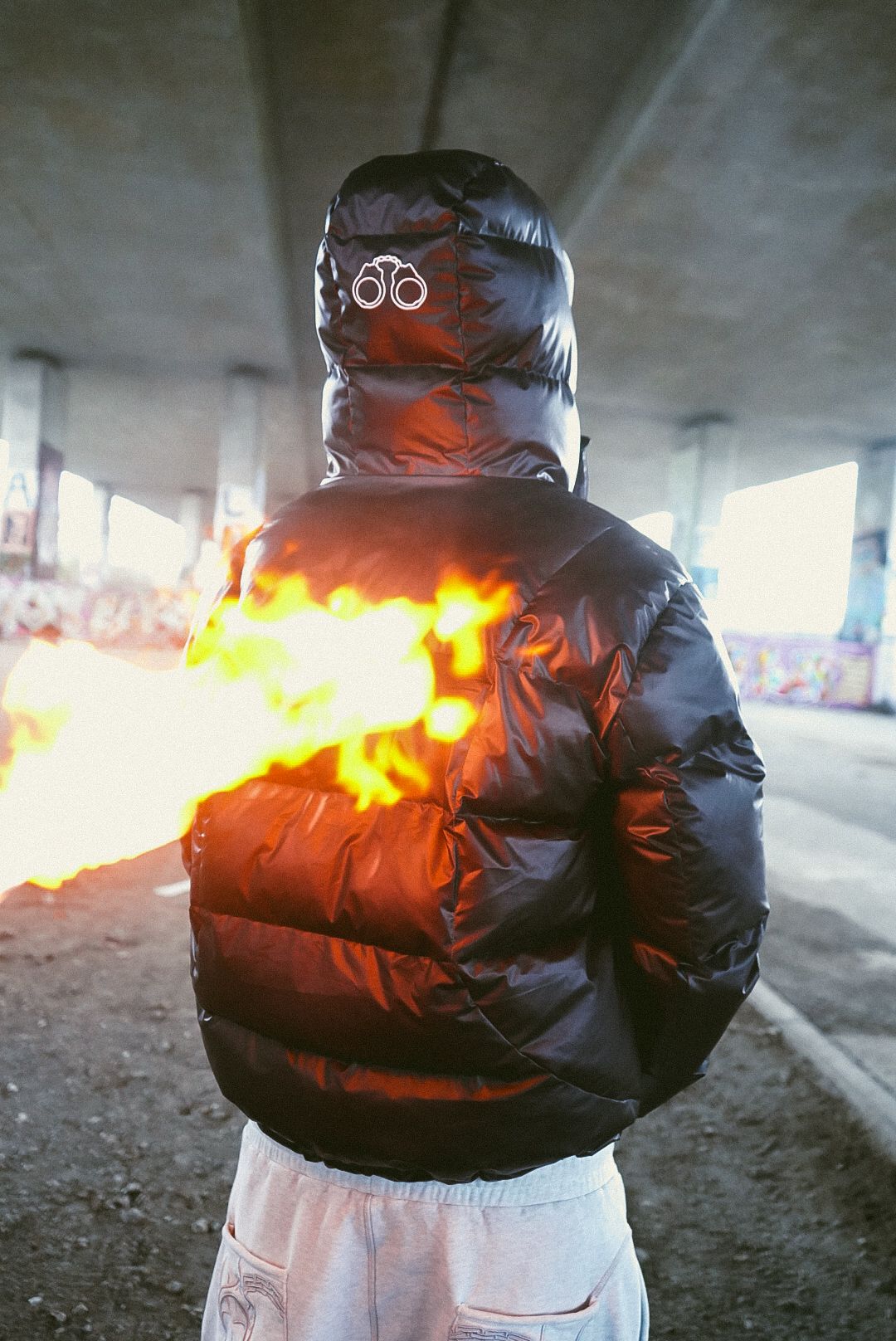 Person in black puffer jacket with hood up, standing with fire effect behind them under bridge.