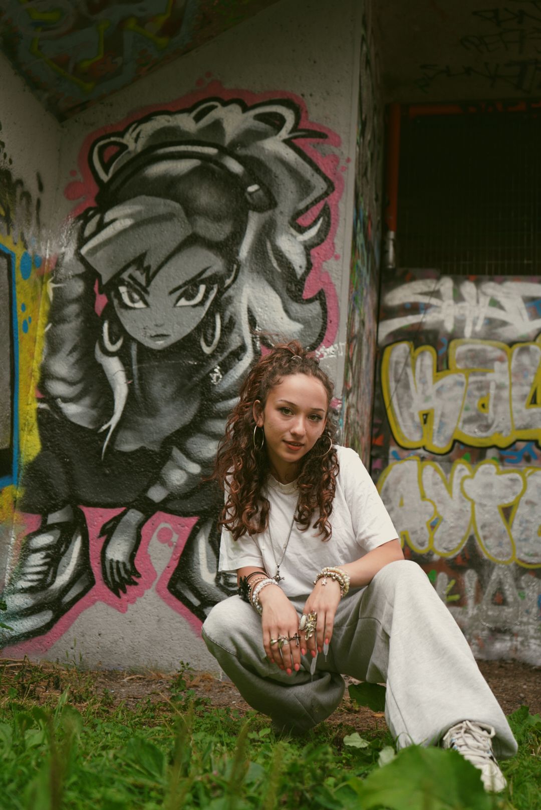 Young woman in white tee and grey pants crouching in front of colorful graffiti mural outdoors.