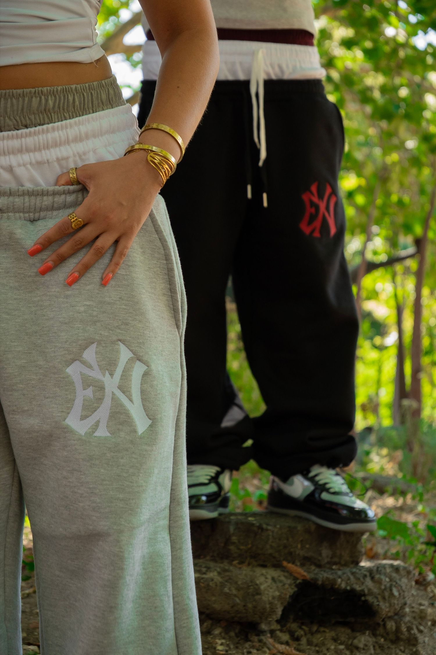 Two people wearing NY logo sweatpants, one gray and one black, standing outdoors among green trees.