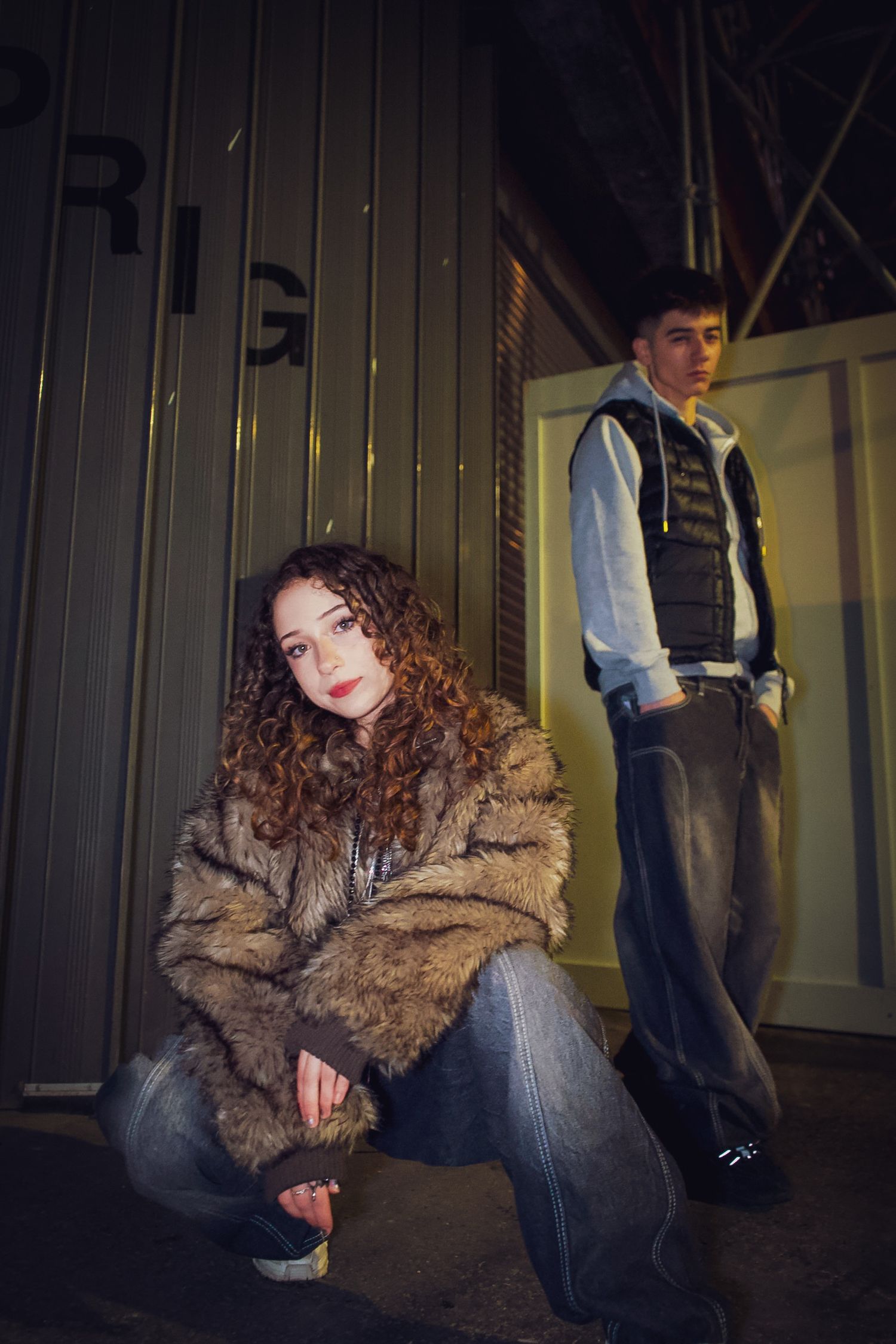 Young woman in fur coat crouching and man in vest standing by metal container at night.