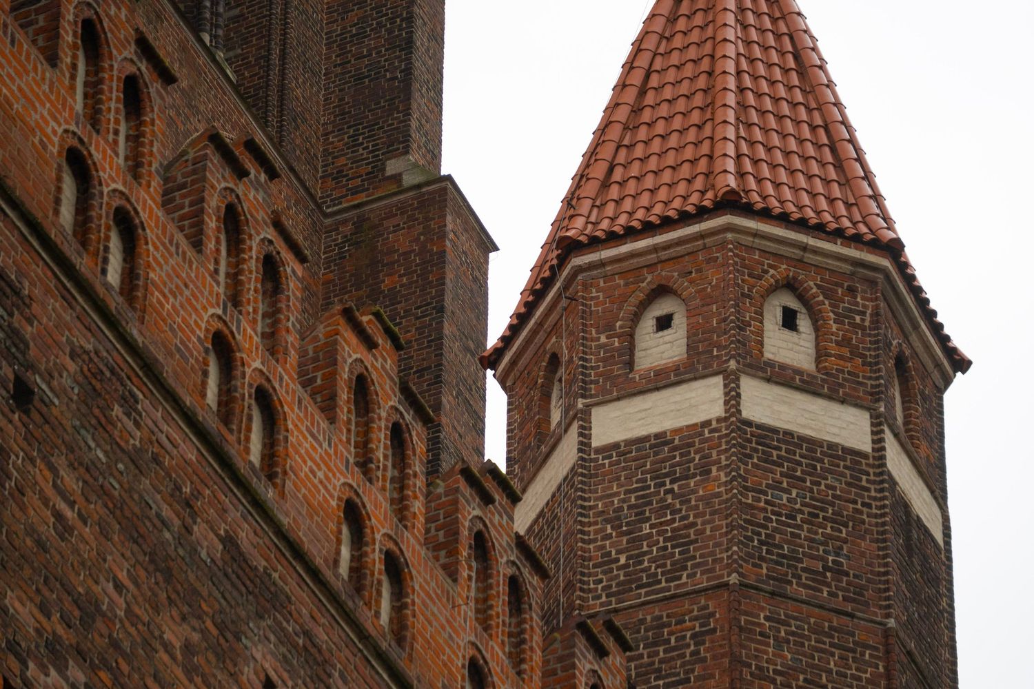 Historic red brick Gothic church tower with terracotta tiled spire roof against overcast sky.