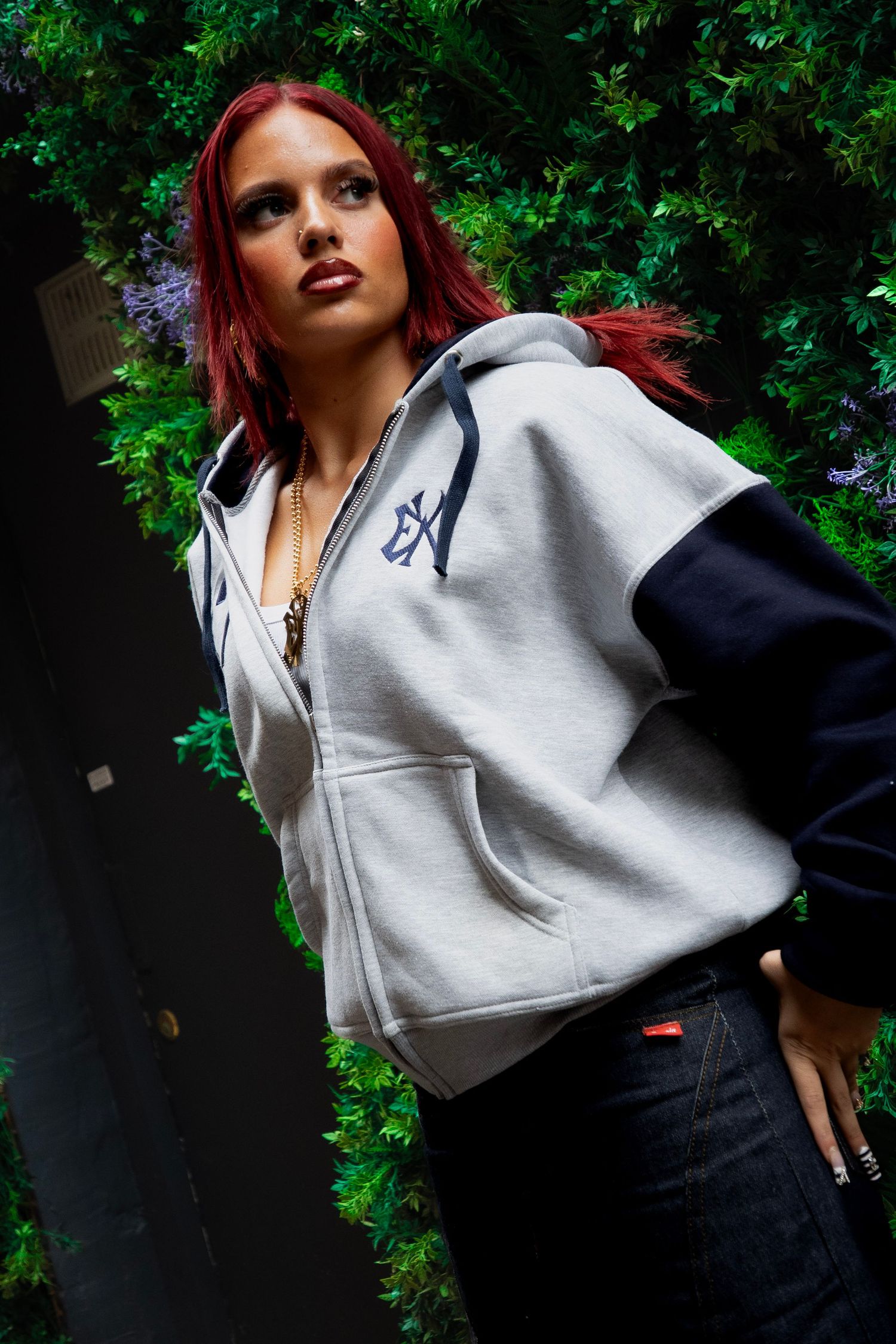 Red-haired woman in gray and navy zip-up hoodie posing confidently against a lush green ivy wall backdrop.