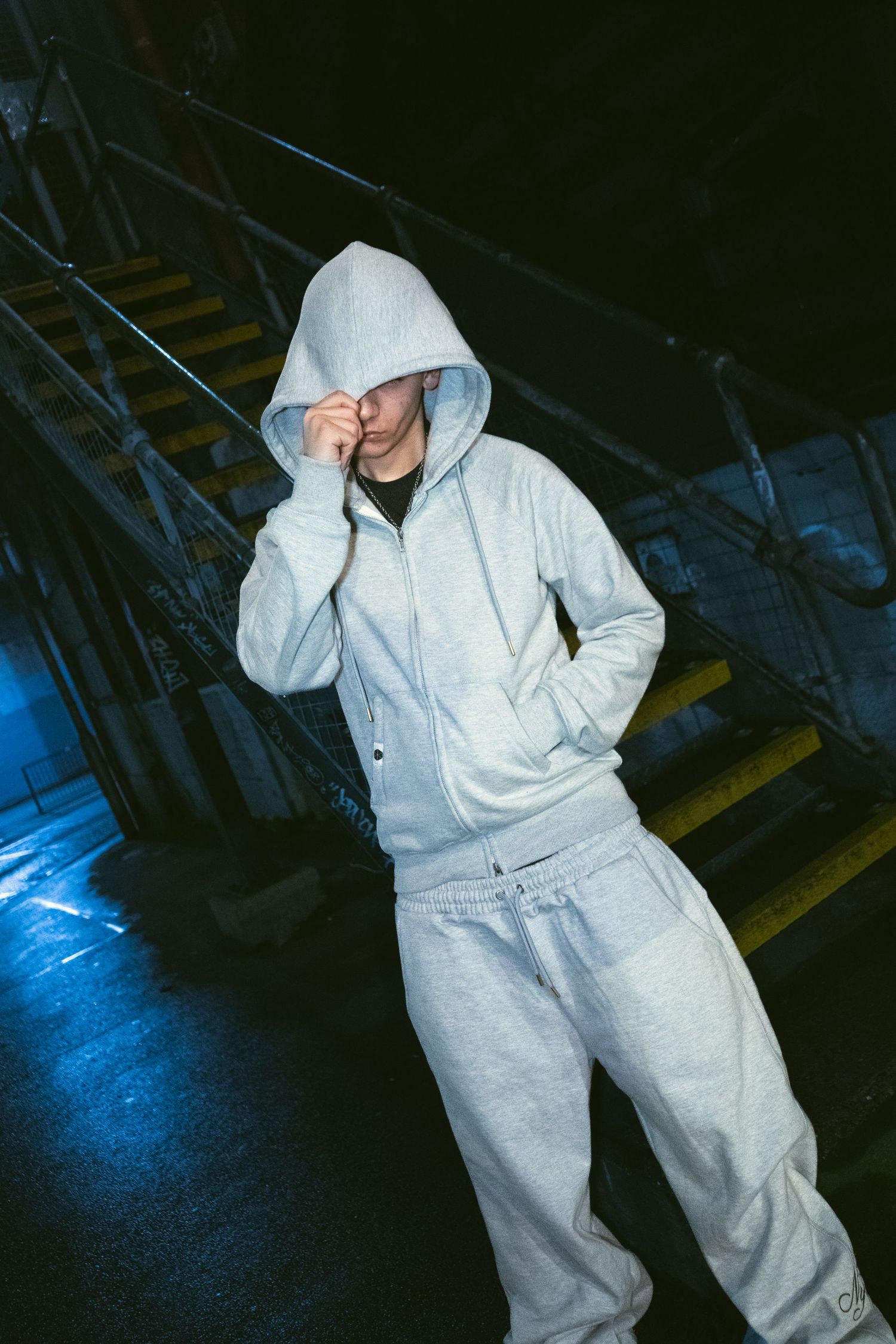 Young man in grey zip-up hoodie and sweatpants posing near industrial staircase in blue light.