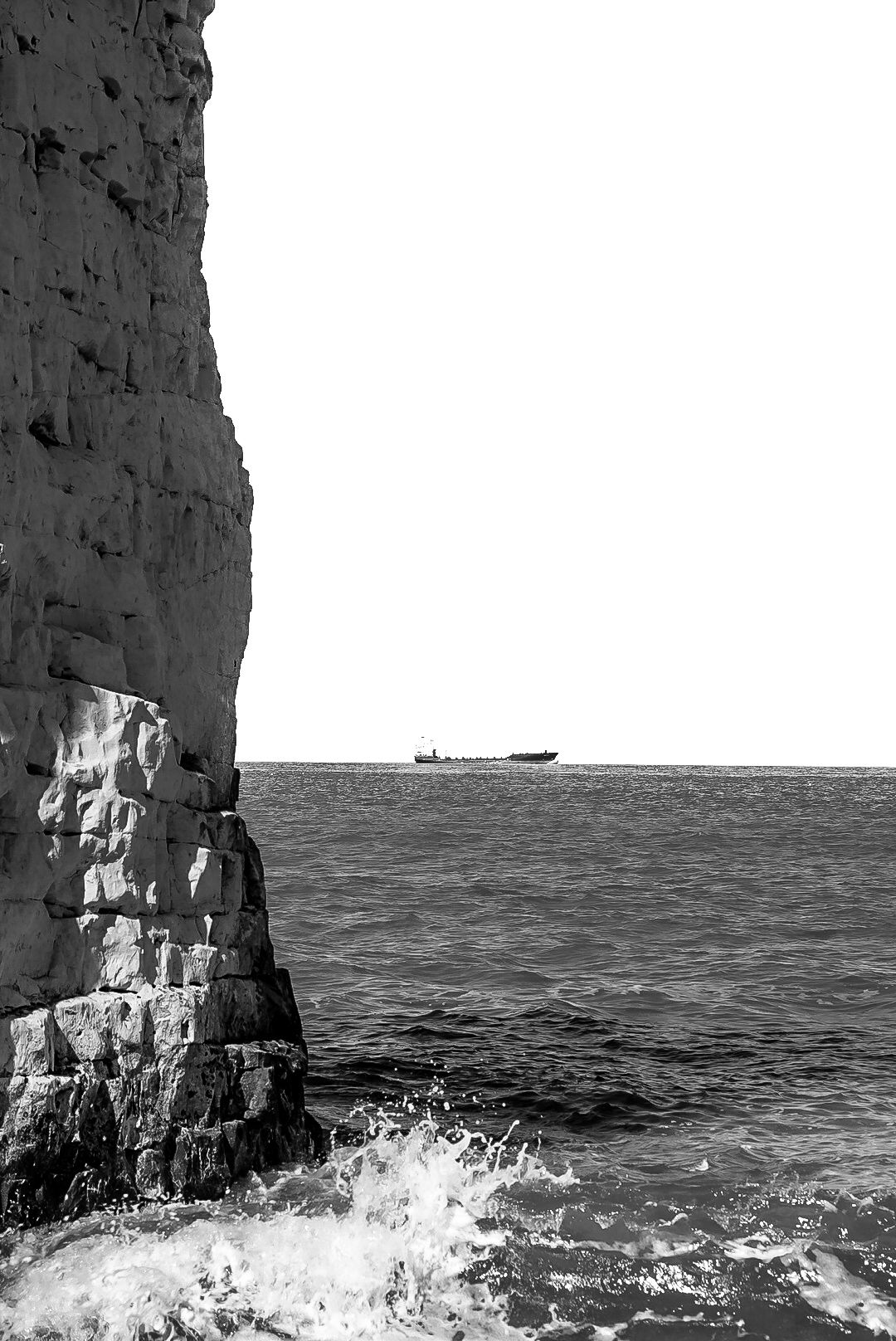 Black and white coastal scene with rugged cliffs, ocean waves, and a distant cargo ship on the horizon.