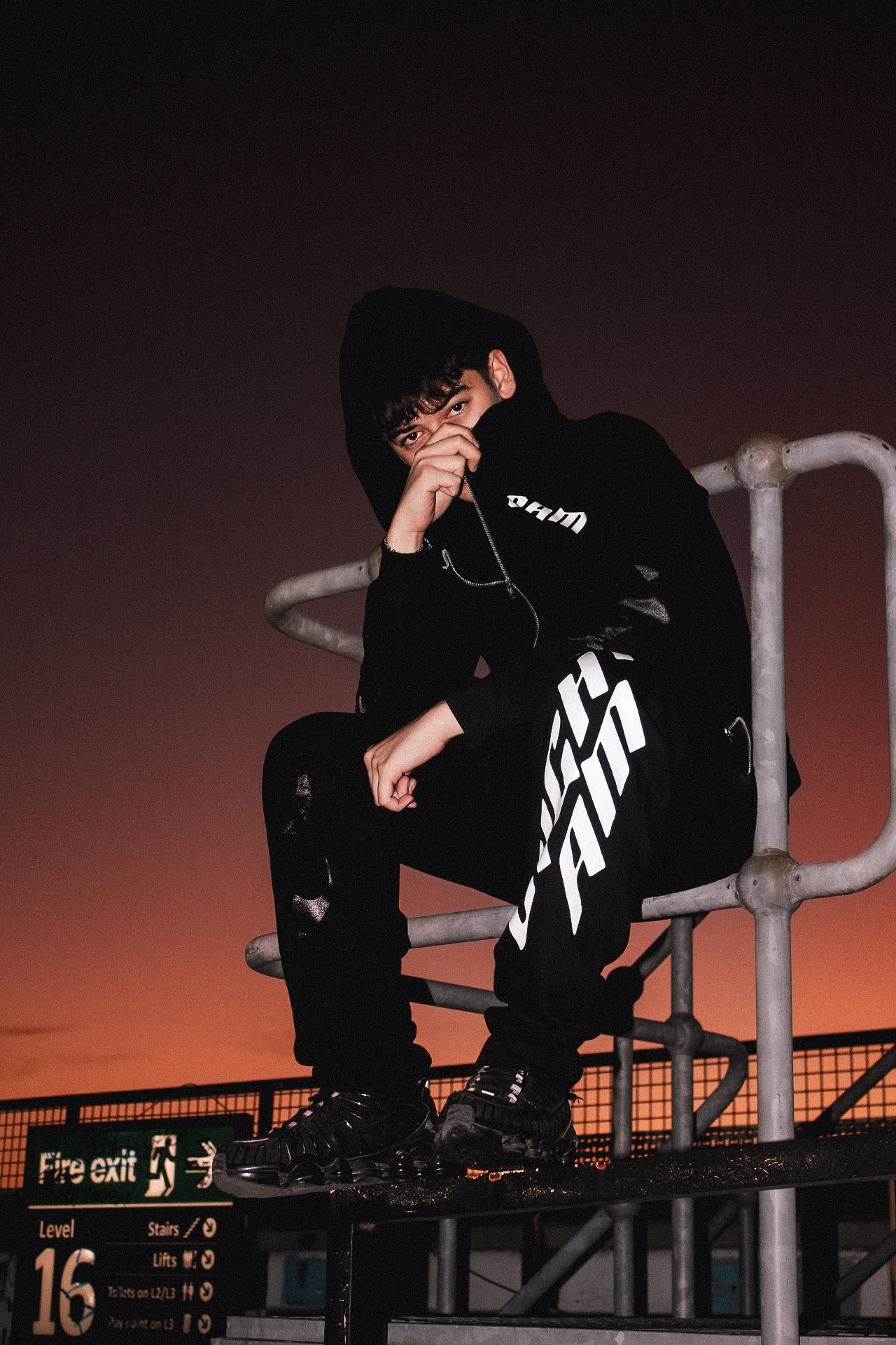 Young man in black graphic hoodie sits on metal railings at sunset with dramatic orange sky backdrop.