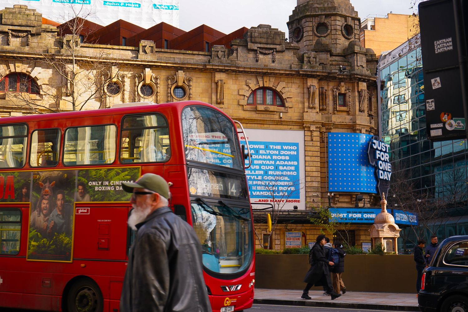 A red double-decker bus passes a historic London theatre on a busy city street.