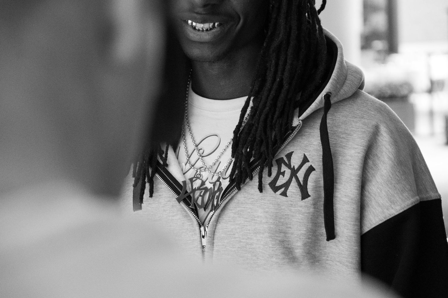 Young man with dreadlocks smiling, wearing a gray and black hoodie with TEX text, black and white photo.