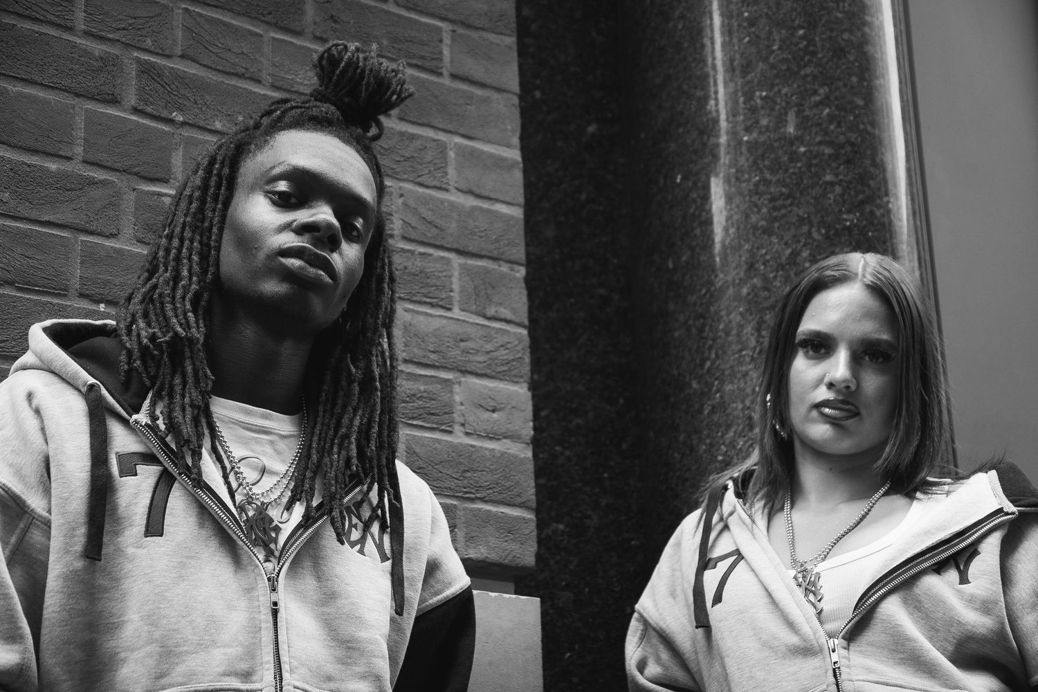 Two young people in matching gray hoodies pose outdoors against a brick wall in a black and white photo.