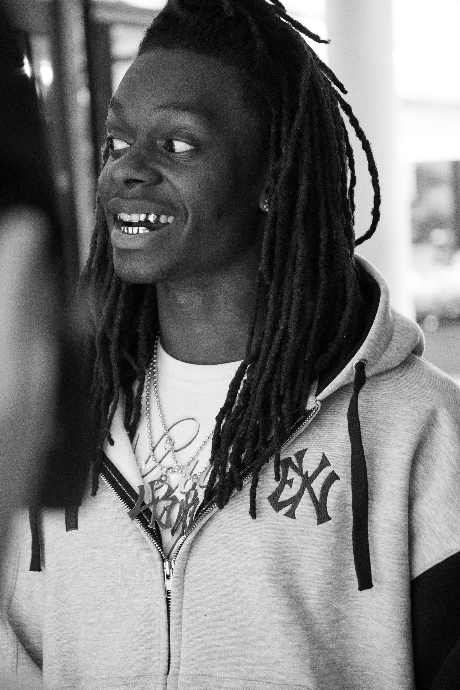 Black and white portrait of a young man with dreadlocks, smiling, wearing a gray zip-up hoodie.